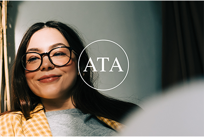 Smiling woman with glasses and dark hair, wearing a yellow checkered shirt, in an indoor setting with a logo reading 'ATA' in the background.