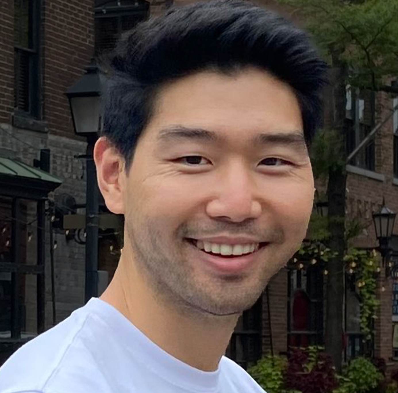 Close-up of a smiling man with dark hair, wearing a white shirt, outdoor urban setting with brick buildings and greenery in the background.