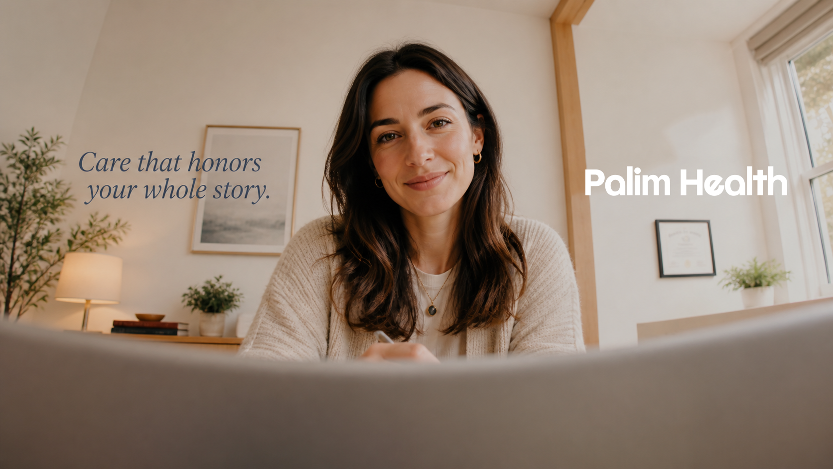 A woman with long dark hair smiling at the camera, sitting in a cozy room with a picture on the wall, plants, and a window, with text overlay reading 'Care that honors your whole story.' and 'Palm Health'.