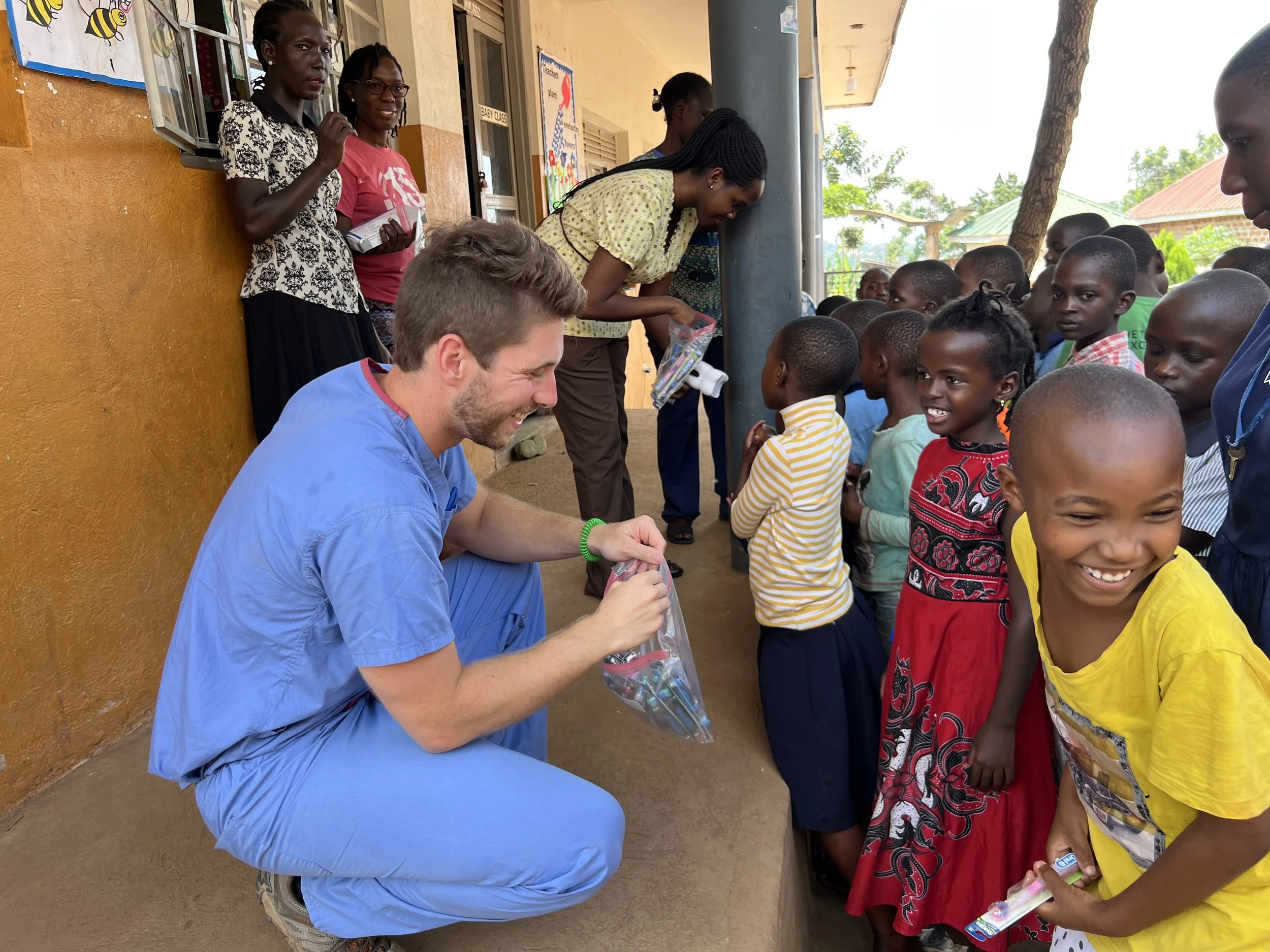 A man in a blue medical uniform sits on a stool handing out toothbrushes to children in line outside a school building.