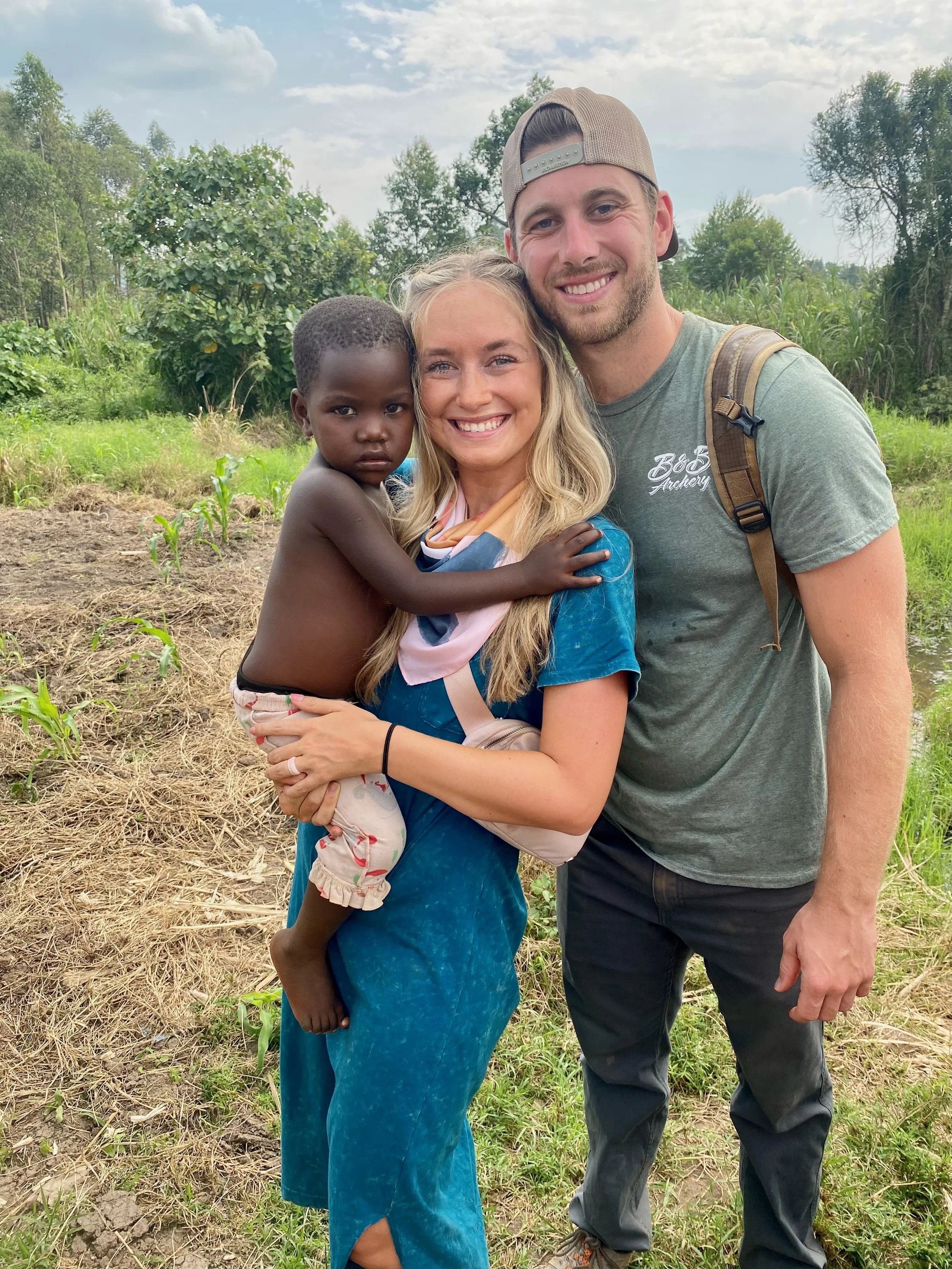 A group of three people outdoors in a field with green plants and trees in the background. A woman with long blonde hair is holding a young child, and a man wearing a baseball cap stands next to them, all smiling at the camera.
