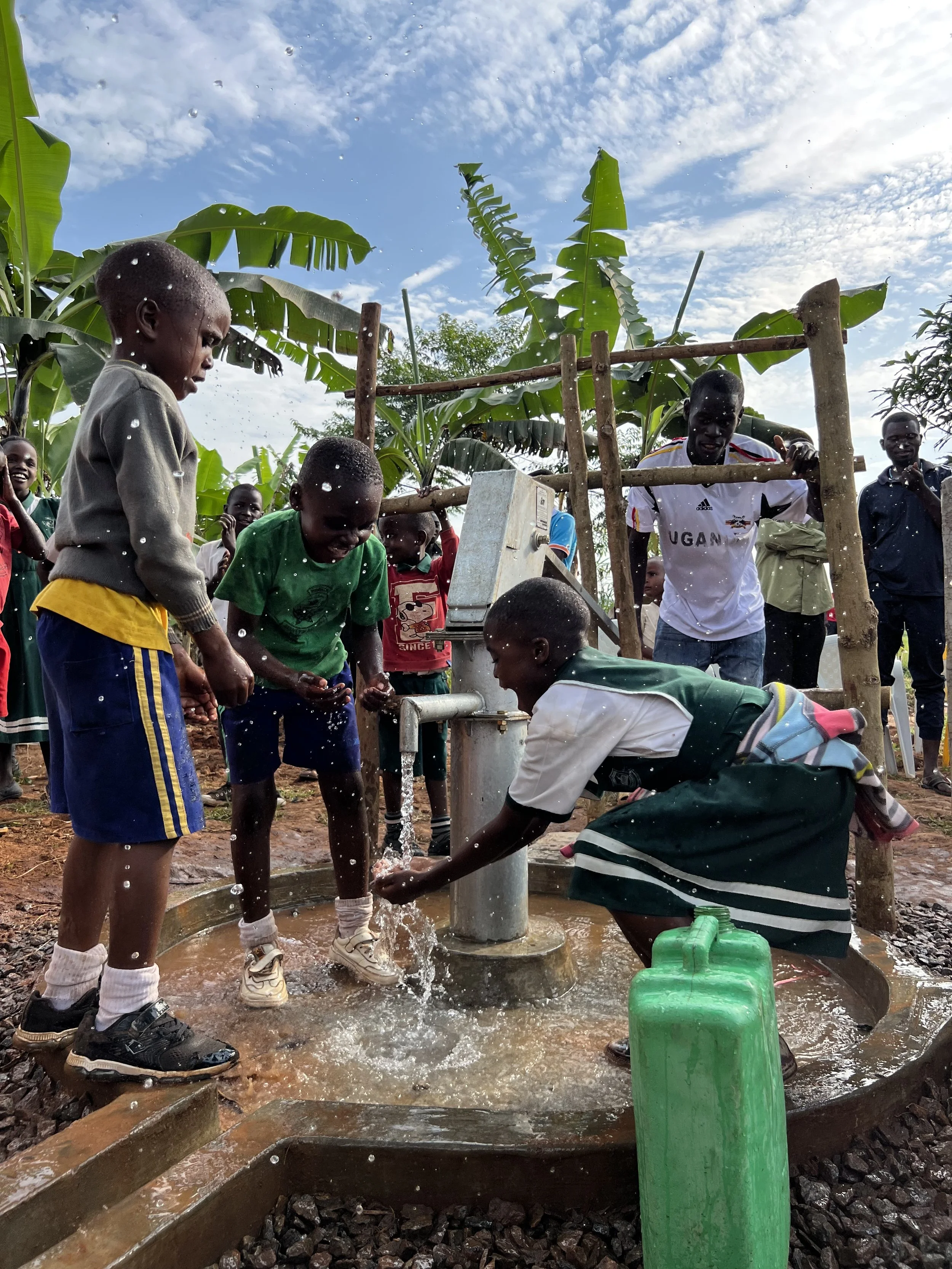 Children and a man collecting water from a hand pump in a rural setting with banana trees and a partially cloudy sky in the background.