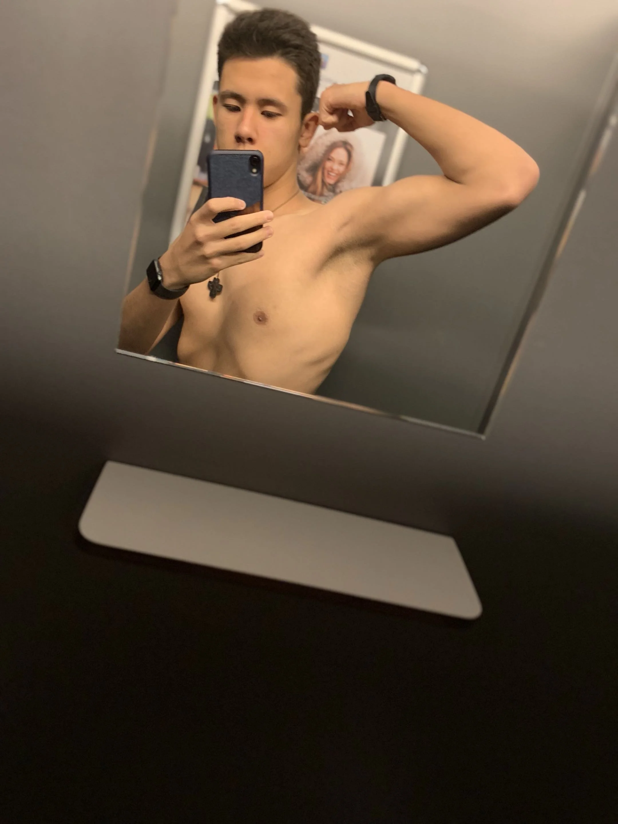 A shirtless young man taking a mirror selfie, flexing his bicep, with a photo of a woman in the background.