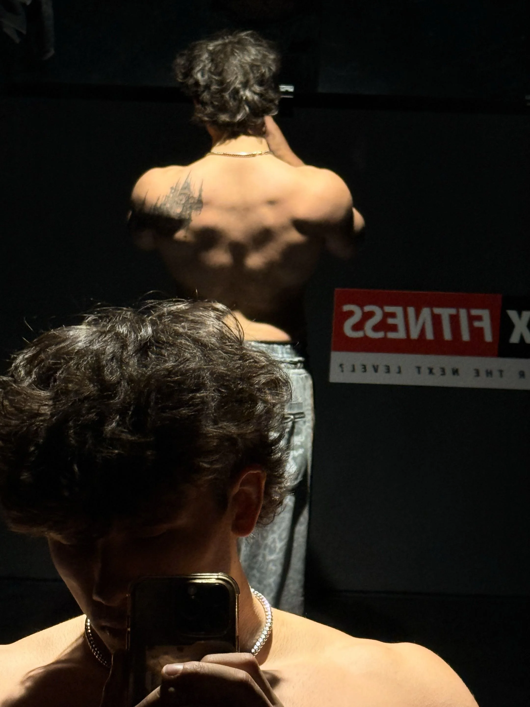 A person taking a mirror selfie in a dimly lit setting, showcasing their short curly hair, tattoos on the back, and wearing a necklace and earrings, with a sign reading 'X Fitness' visible in the background.