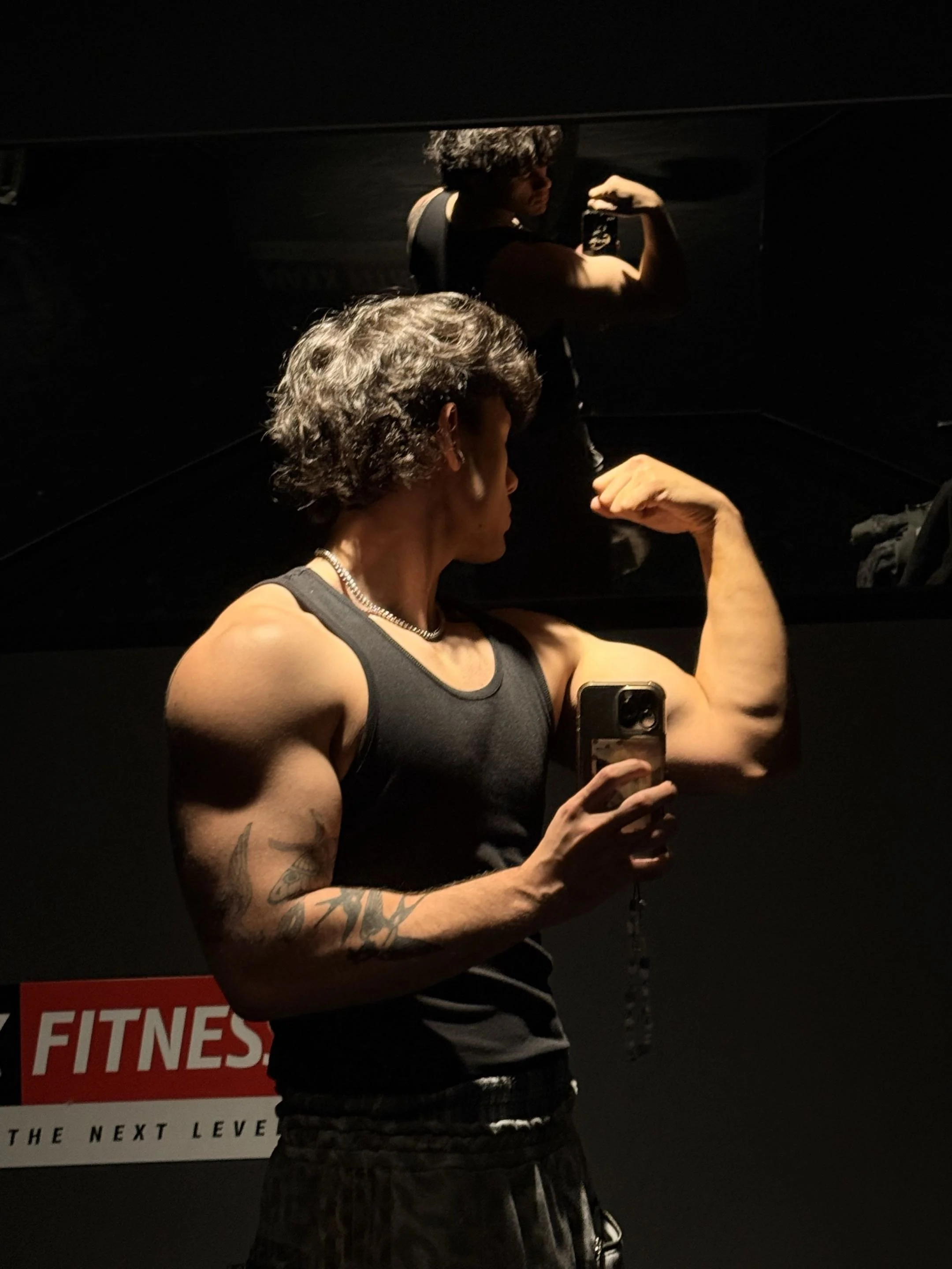 A muscular man taking a selfie in a dark gym, flexing his bicep, with a mirror reflecting his image and a sign about fitness in the background.