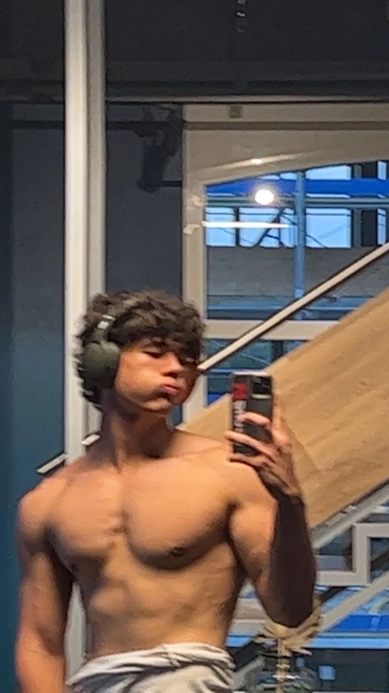 A shirtless young man with curly hair taking a mirror selfie in a room with large windows and a staircase.