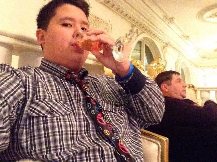 A young person in a plaid shirt drinking from a glass with a slice of pizza, sitting in a lavishly decorated room with ornate moldings and gold accents, with a man in the background sitting at a table.