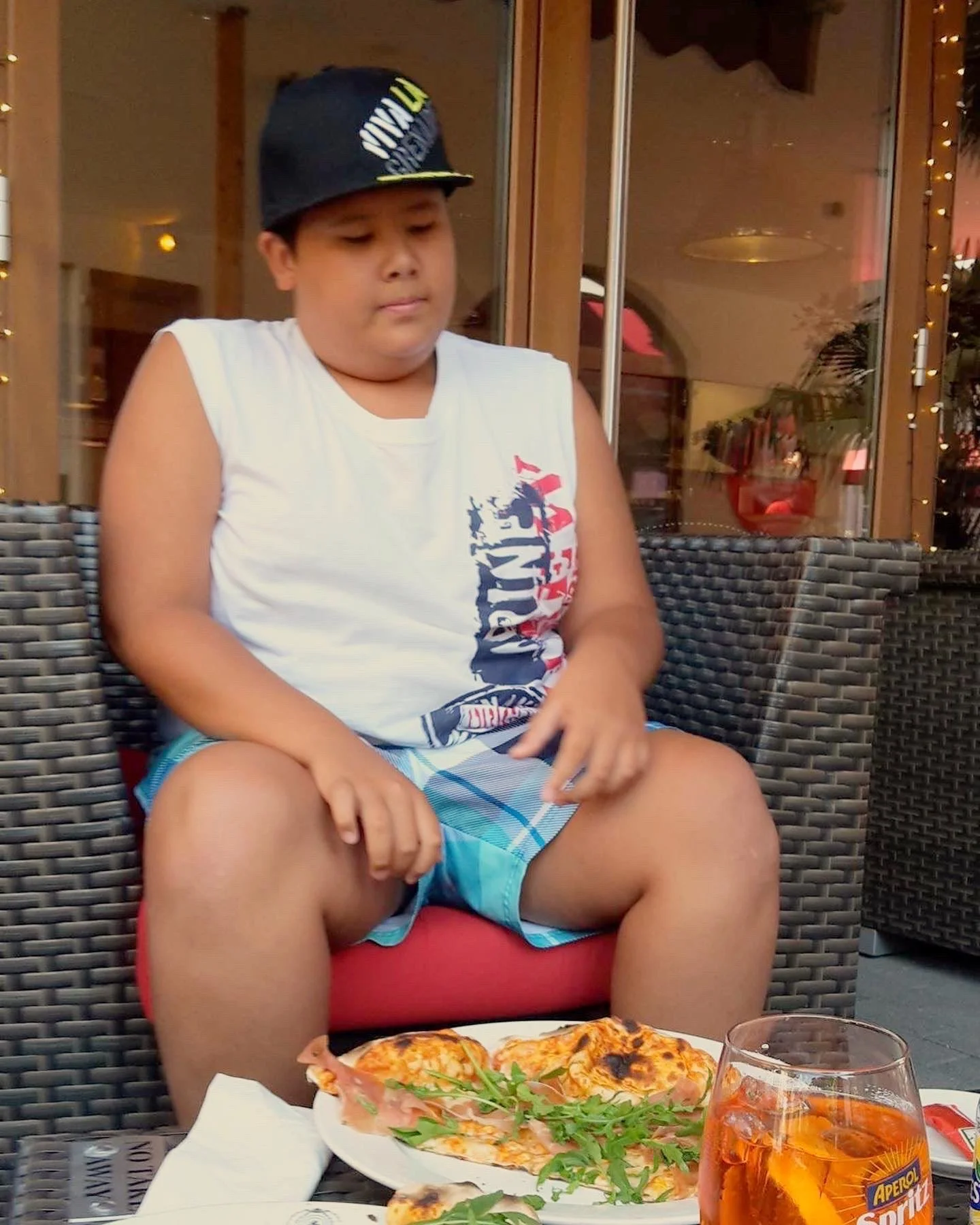 A boy sitting outdoors at a restaurant table with pizza and a drink. The boy is wearing a sleeveless shirt, plaid shorts, and a baseball cap. There are wicker chairs, a wooden structure, and lights in the background.
