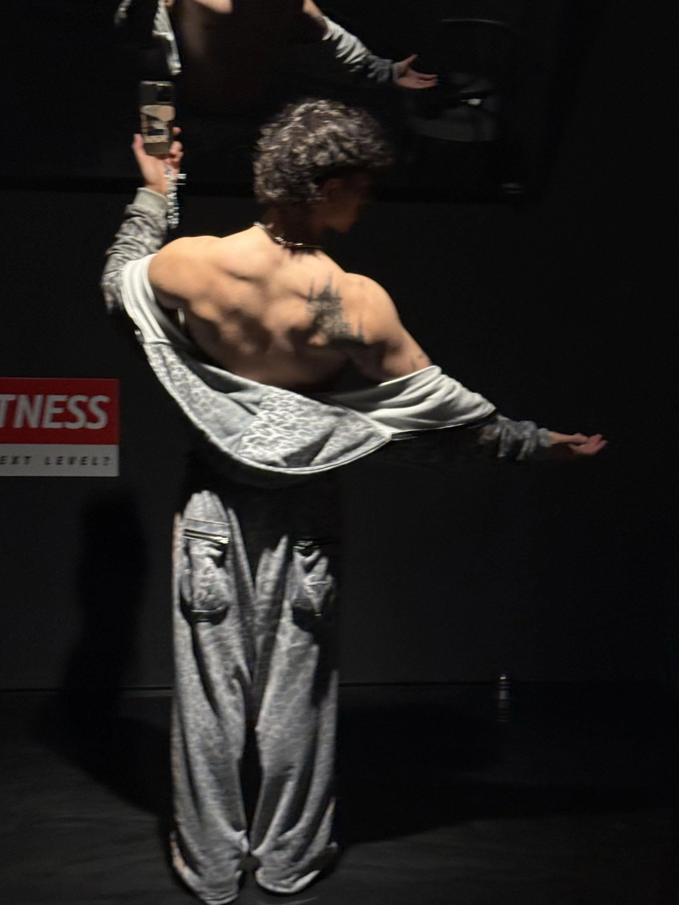 A person with curly gray hair taking a mirror selfie in a dark room, wearing shiny silver and black patterned clothing with a sleeveless top and loose pants, background is black with a partial red and white sign.