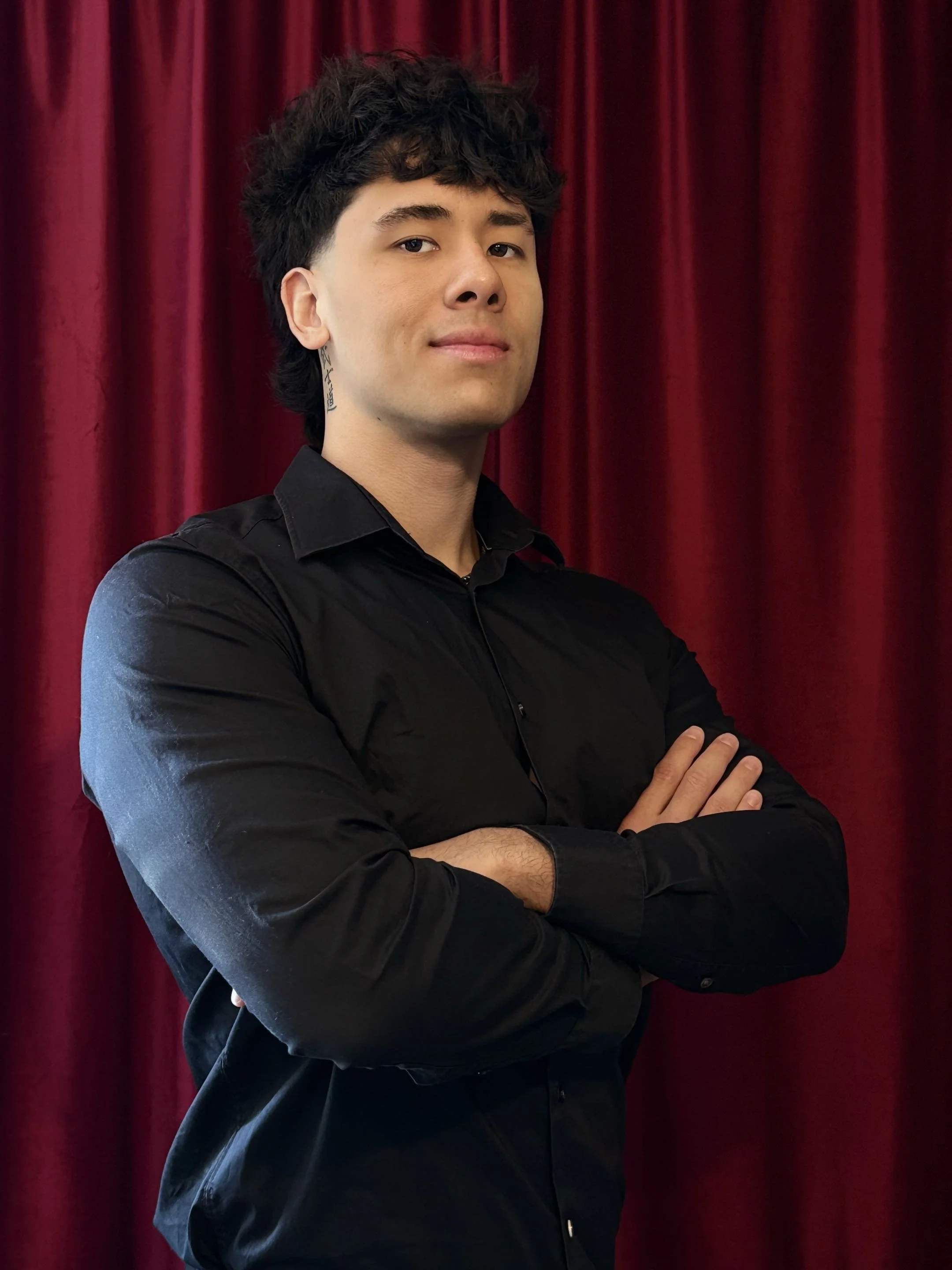 A young man with dark, curly hair posing with arms crossed in front of red velvet curtains, wearing a black shirt.