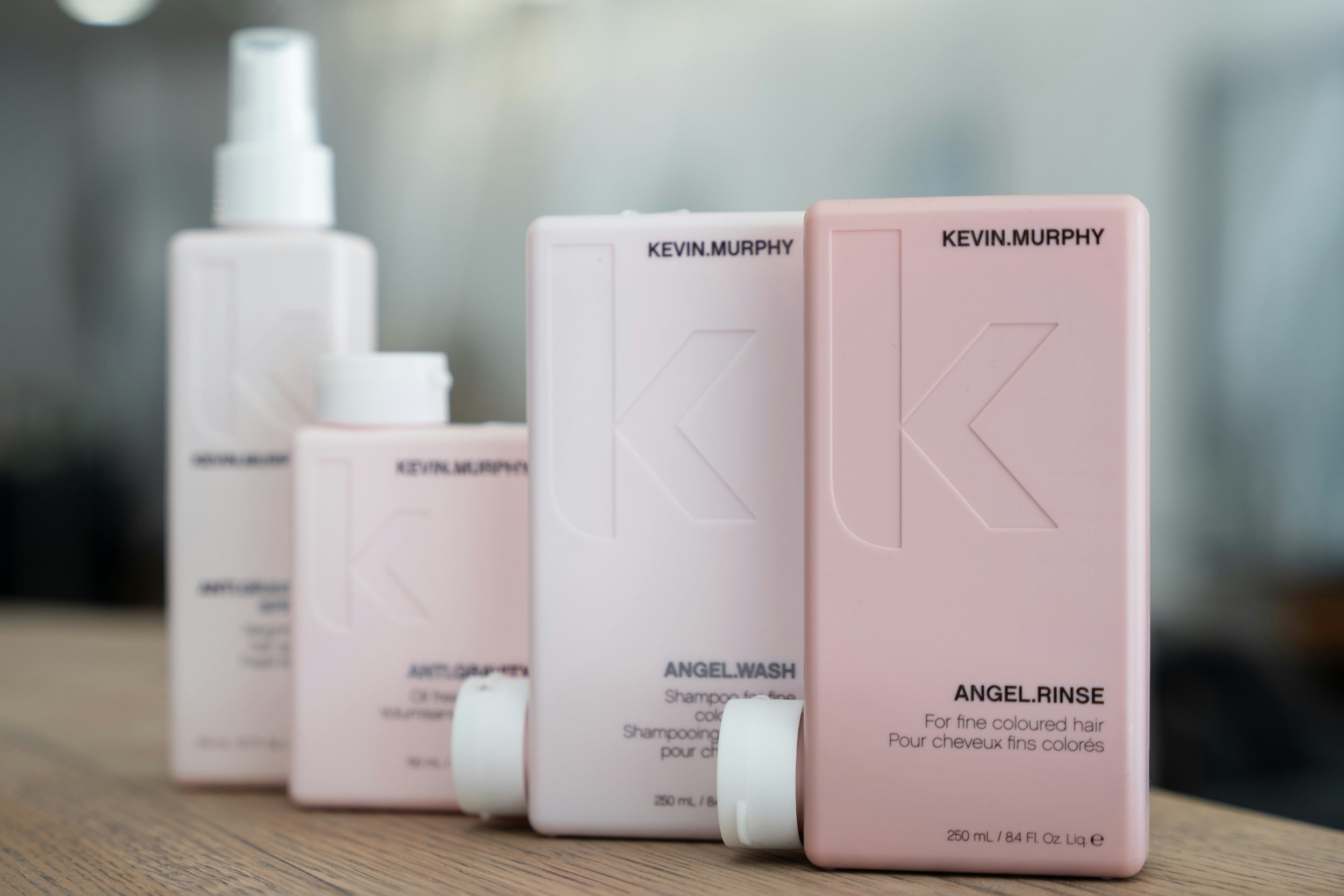 Rows of pink and white hair care product bottles with labels, on a wooden surface. The labels include the brand name 'KEVIN.MURPHY' and product descriptions like 'ANGEL WASH' and 'ANGEL RINSE'.