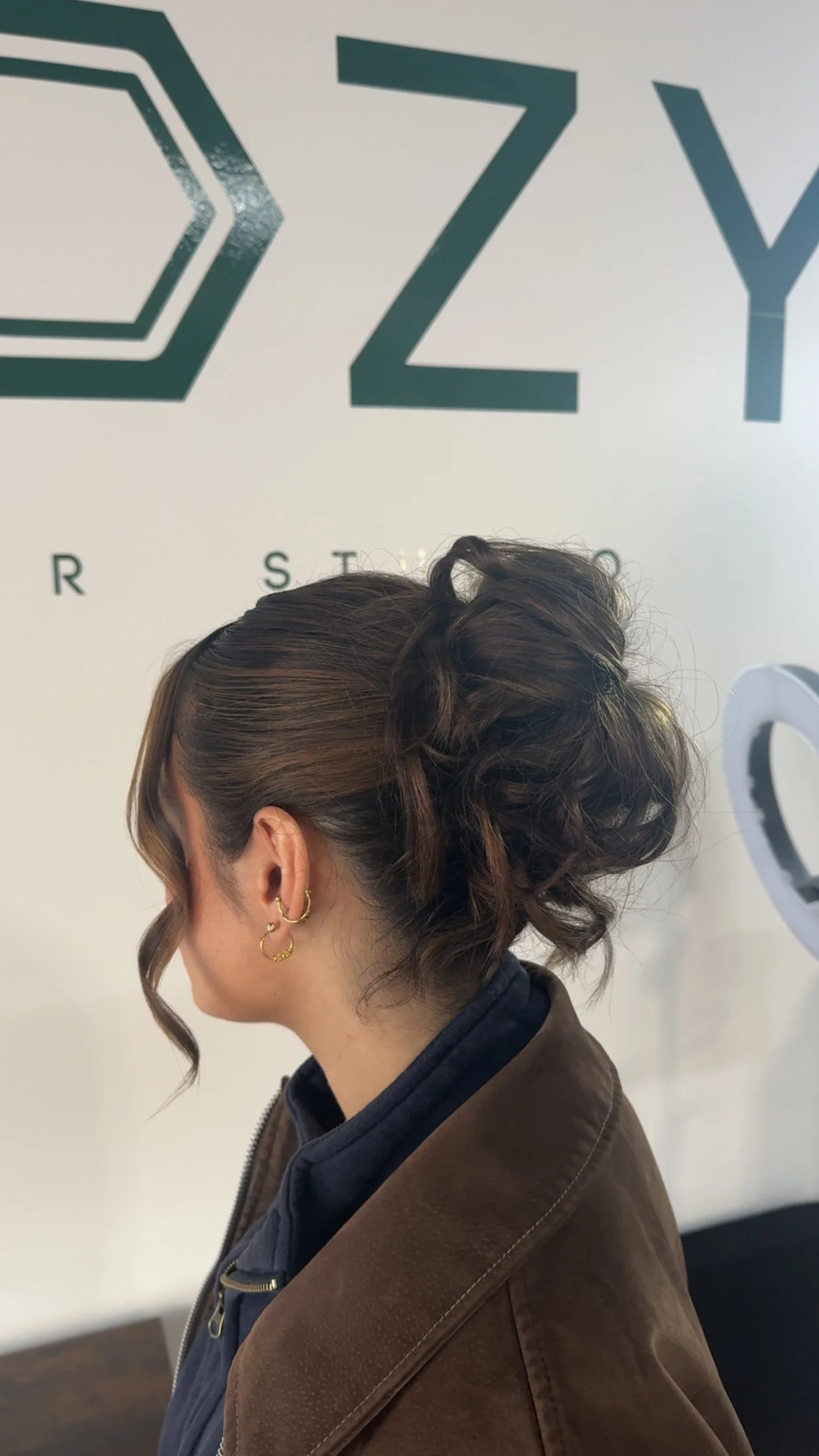 A woman with brown hair styled in an elegant updo, wearing gold earrings and a brown jacket, standing in front of a white wall with the partial logo text 'OZY' in green.