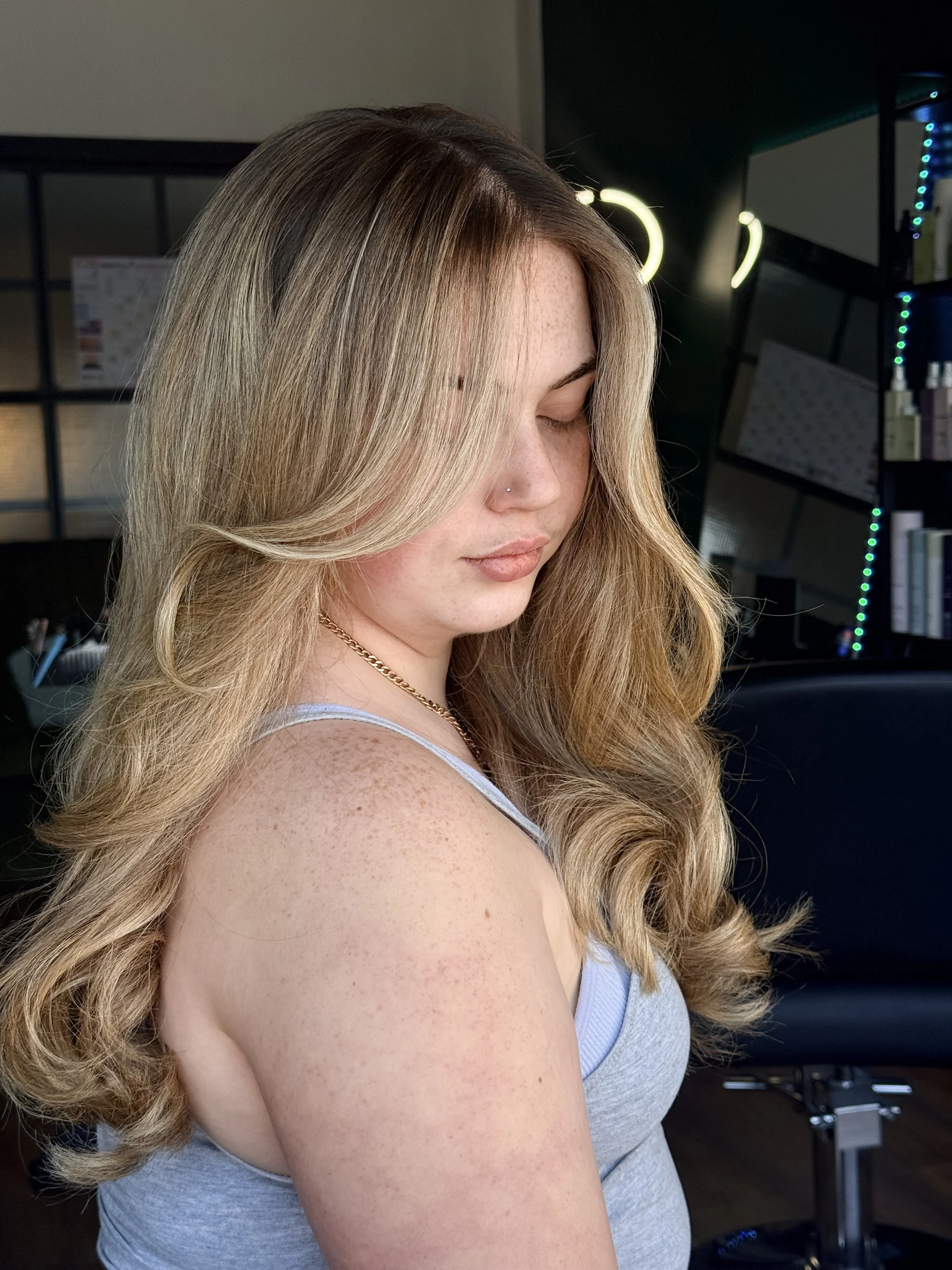 A woman with long, wavy blonde hair and fair skin is in a side profile, with her eyes closed, in a modern interior with dark walls, illuminated mirror lights, and shelving.