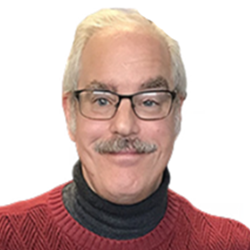 Close-up of a man with gray hair, glasses, a mustache, wearing a red sweater over a black turtleneck, smiling.