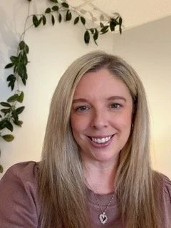 A smiling woman with long blonde hair, wearing a mauve top and a heart-shaped necklace, standing in front of a white wall with a green plant.