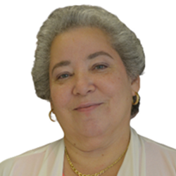 A portrait of an older woman with gray hair, smiling, wearing earrings, a gold necklace, and a light-colored blazer.