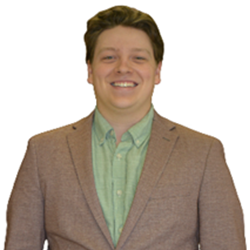 A smiling young man wearing a brown blazer and light green shirt, standing against a plain white background.