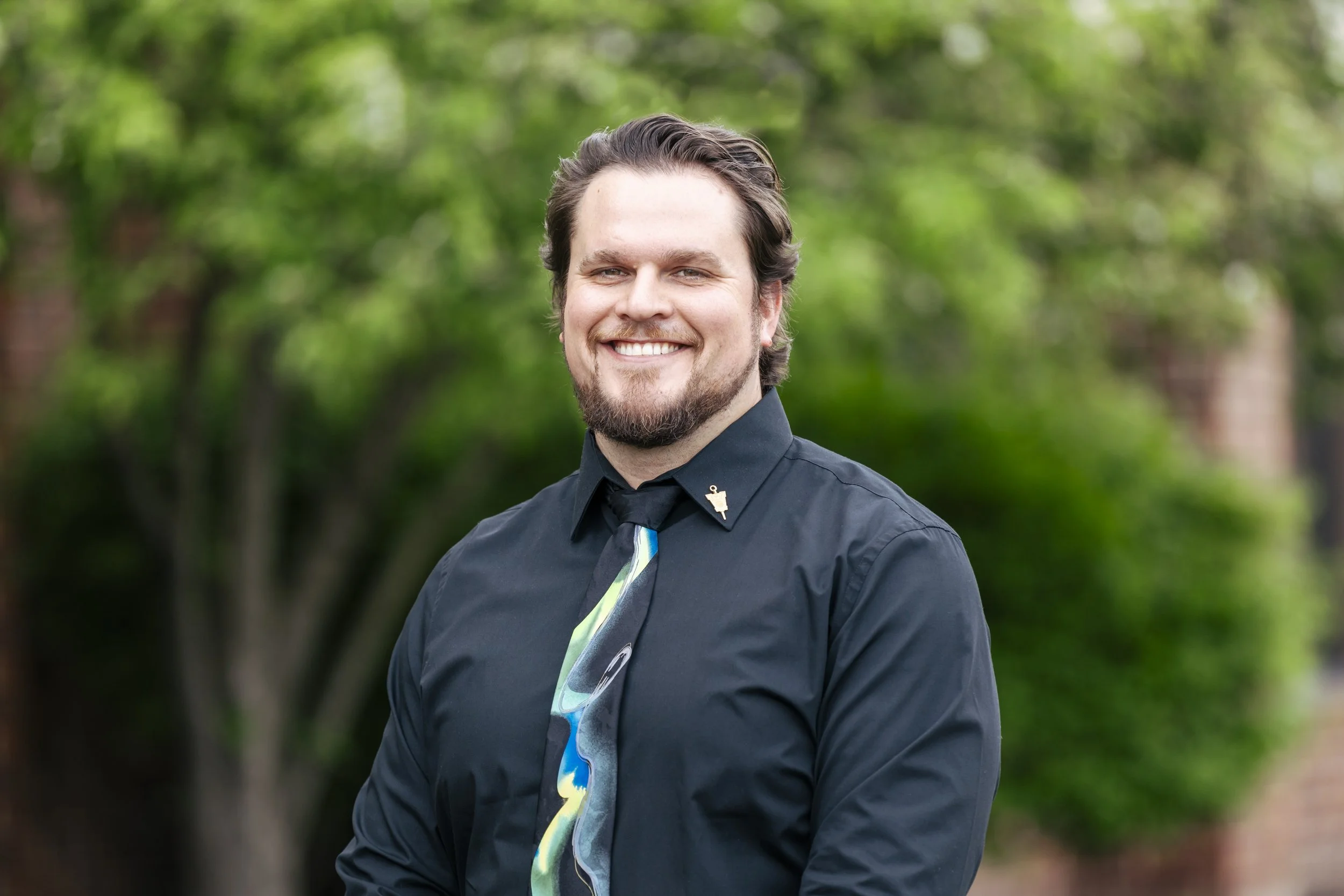 A smiling man with dark hair and a beard dressed in a black shirt and tie, standing outdoors with green trees in the background.