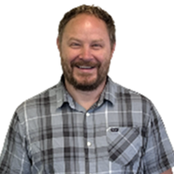 A smiling man with a beard and curly hair wearing a gray plaid shirt.