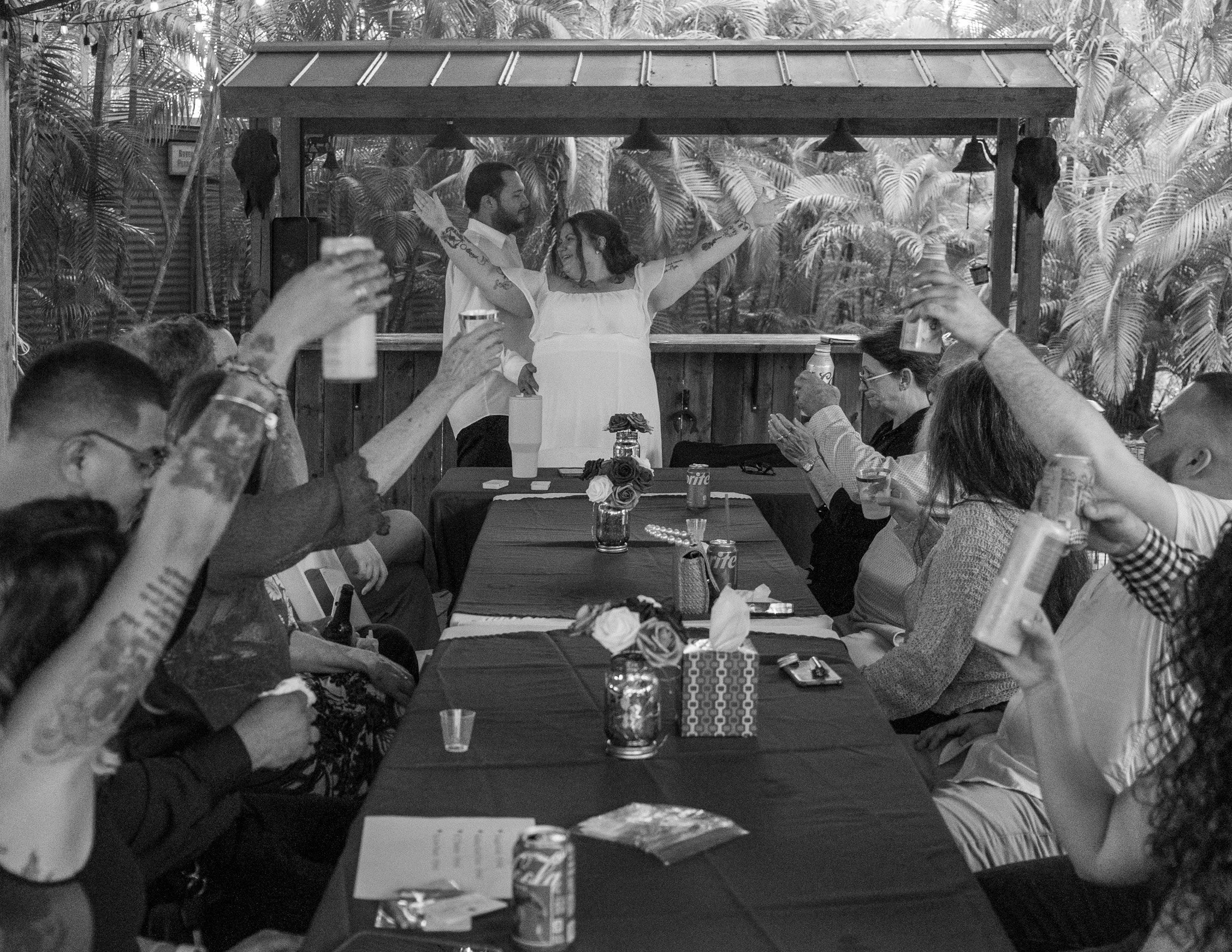 A couple dancing at a celebration with guests sitting at a long table, drinks, flowers, and decorations, with tropical plants in the background.
