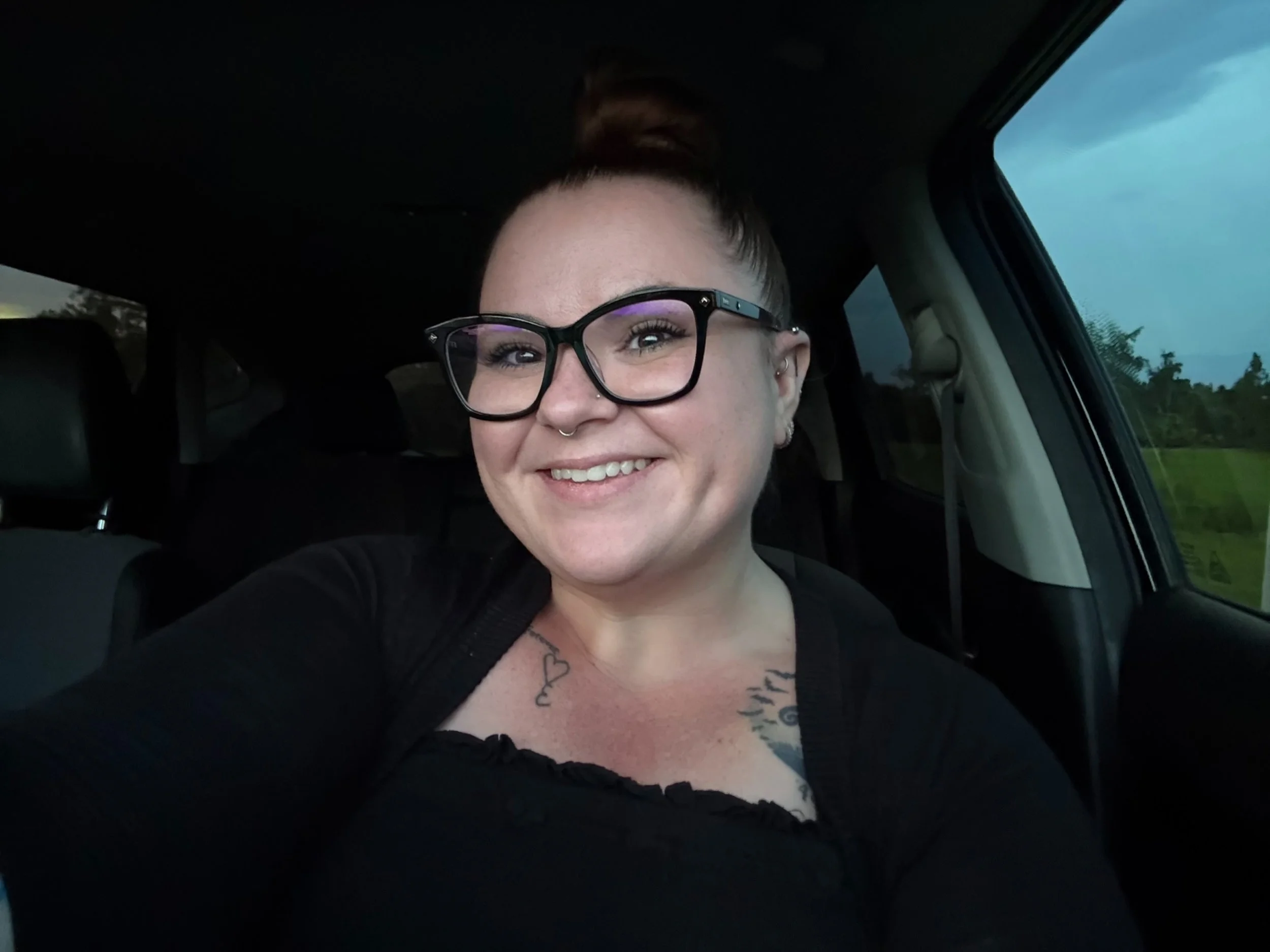 A smiling woman with glasses sitting in a car, with blue sky and trees visible through the window.