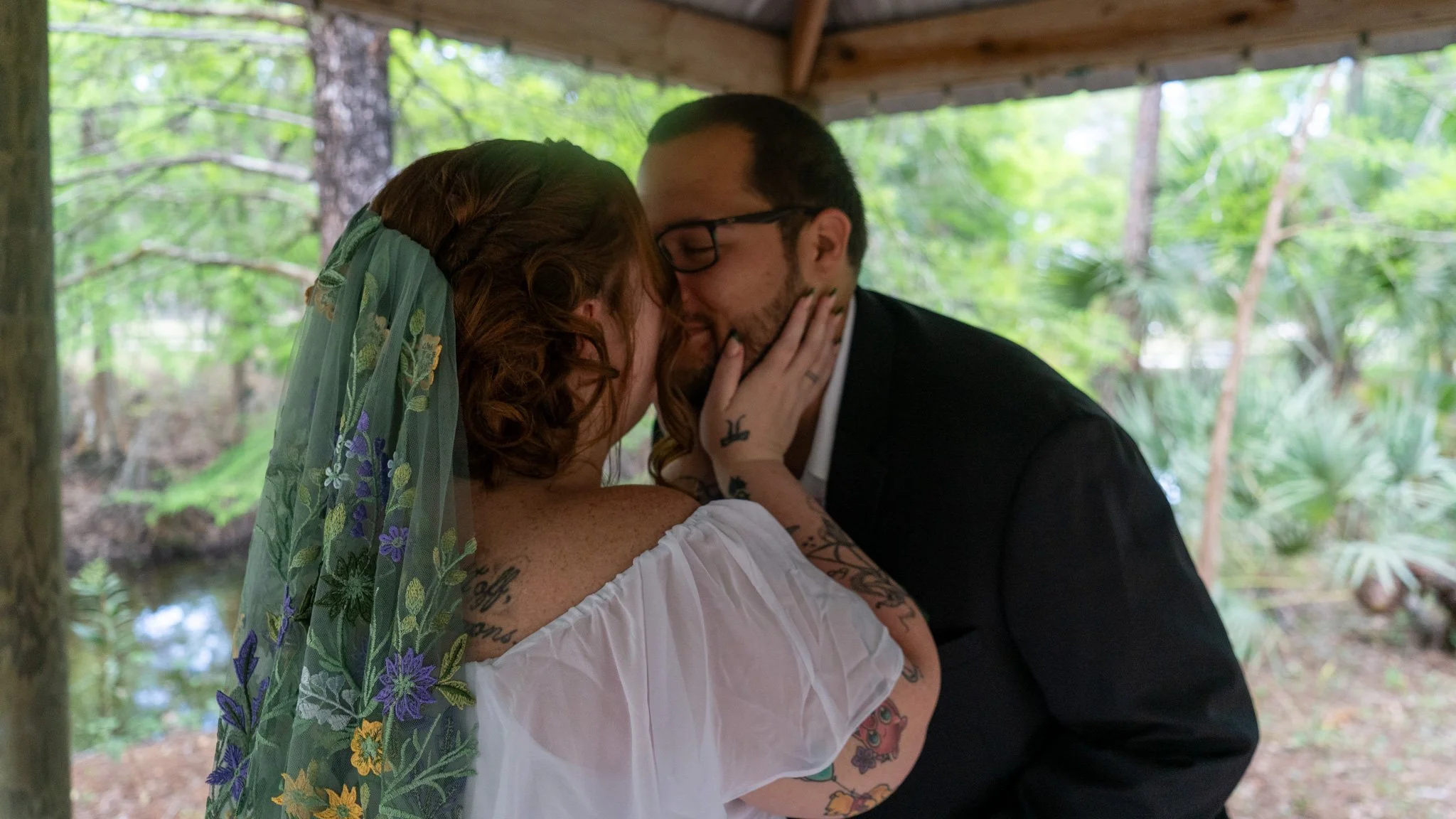 A couple sharing a kiss under a wooden pavilion in a forest, with the woman wearing a veil and tattoos, and the man in a black suit and glasses.