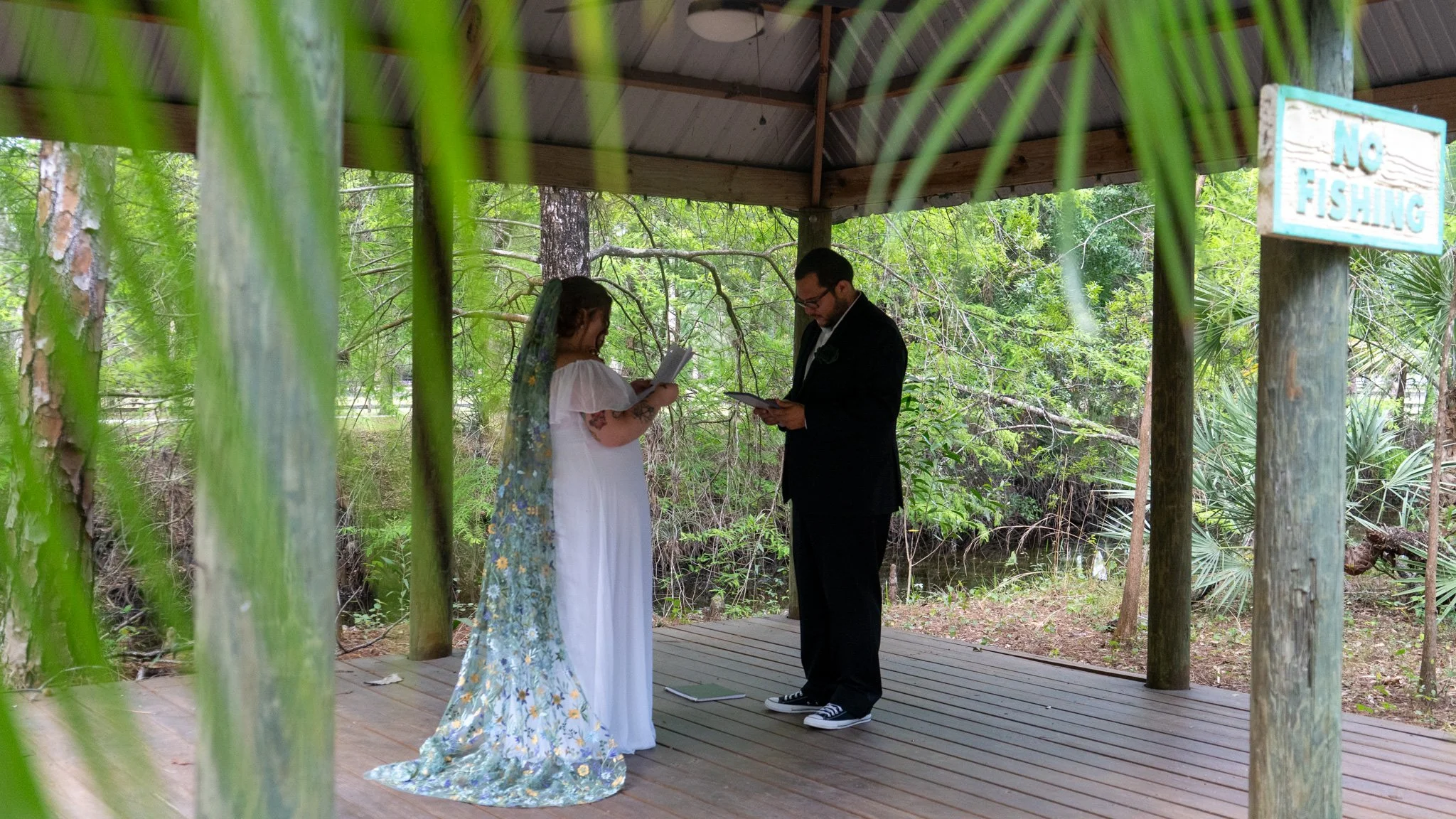 A wedding ceremony taking place outdoors under a gazebo in a wooded area. The bride, in a white dress and a floral veil, is standing with a man in a black suit who is reading from a document. Green foliage is visible around them, and a sign that says 'No Fishing' is attached to a post on the right.