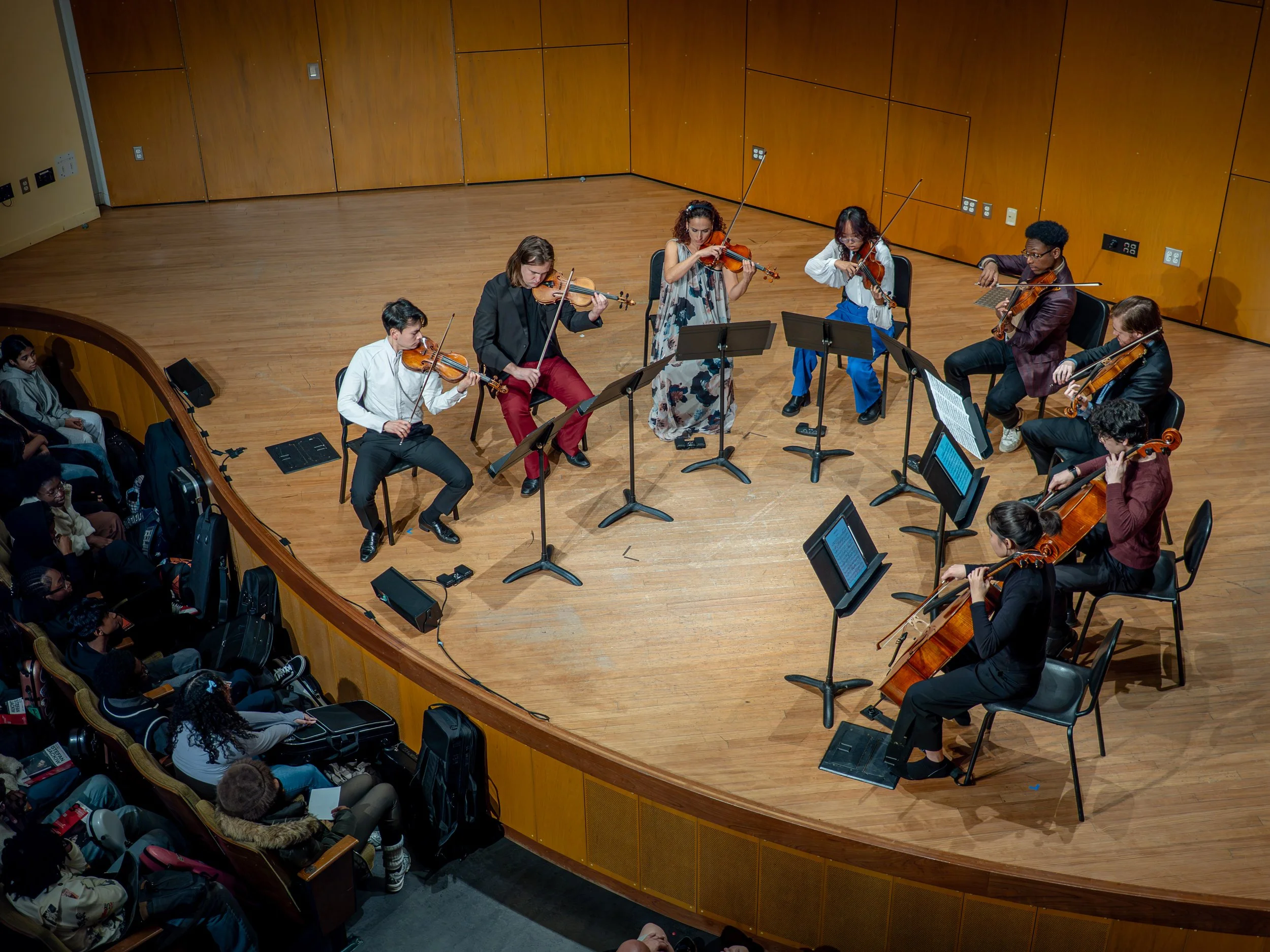Group of musicians performing on stage with string instruments, audience watching in an auditorium.