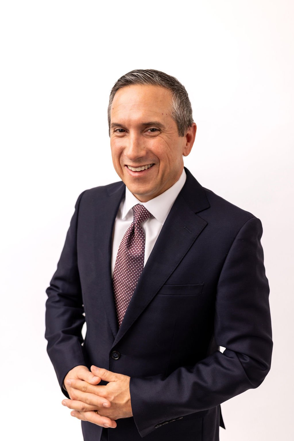 A professional man in a dark suit, white shirt, and maroon patterned tie, standing against a plain white background, smiling with hands clasped in front.
