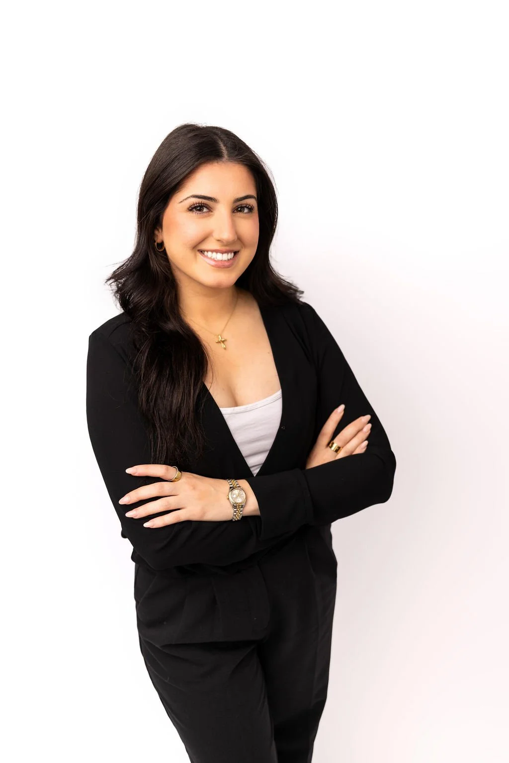 A confident woman with long dark hair smiling, wearing a black blazer over a white top, accessorized with a gold star necklace, rings, and a wristwatch, standing with crossed arms against a plain white background.
