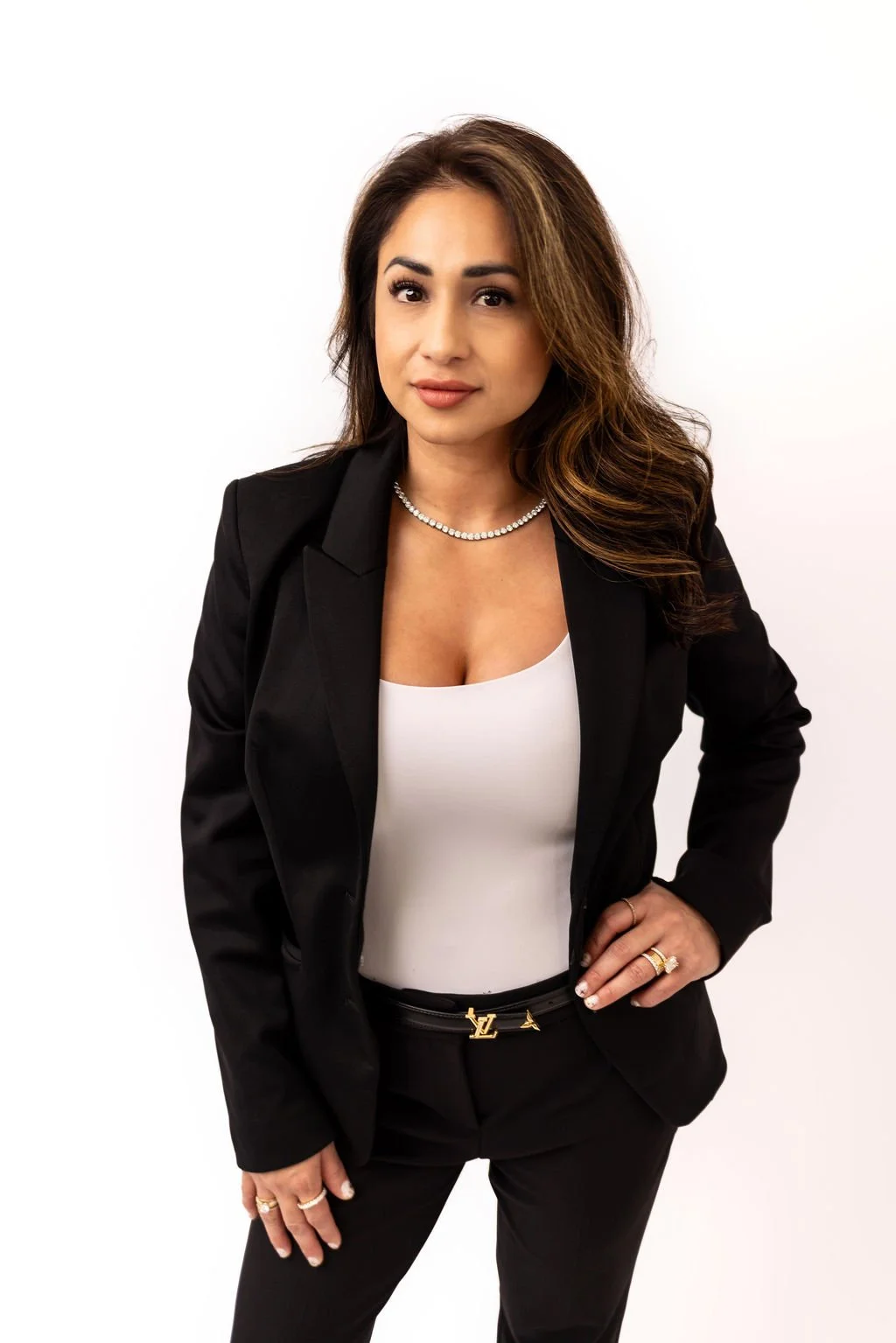 A woman with long brown hair and light makeup, wearing a black blazer over a white top, accessorized with a pearl necklace and rings, standing against a white background.
