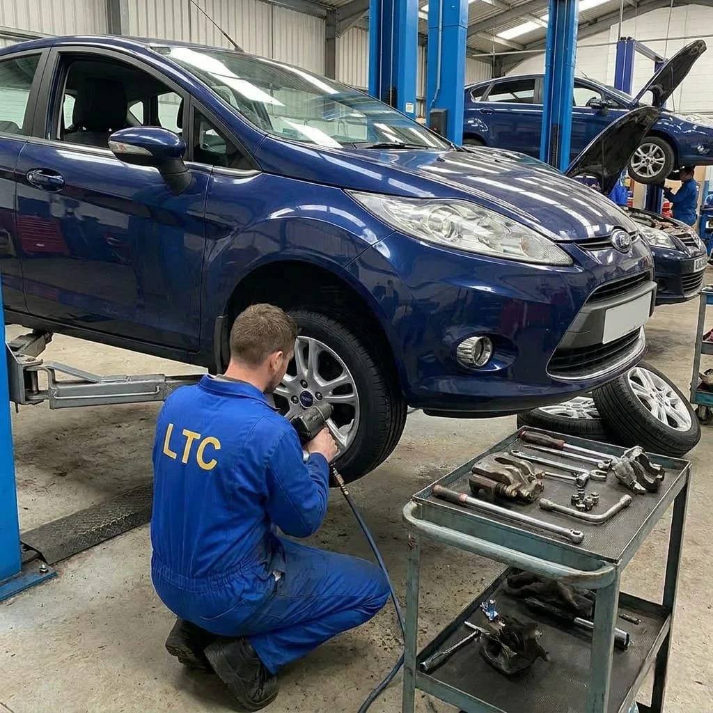 An automotive technician in a blue uniform kneels on the floor, working on the front wheel of a blue car lifted on a hydraulic lift in an auto repair shop. The shop has tools on a nearby metal cart, and there are other cars and mechanics in the background.