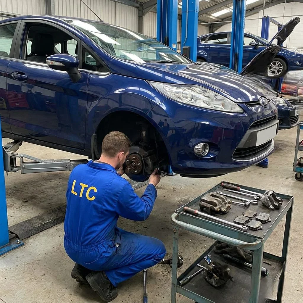 A mechanic working on the front wheel of a blue car lifted on a hydraulic lift inside an auto repair shop. The mechanic is wearing a blue jumpsuit with yellow letters 'LTC' on the back, and there is a metal tool cart nearby with various tools and brake parts.