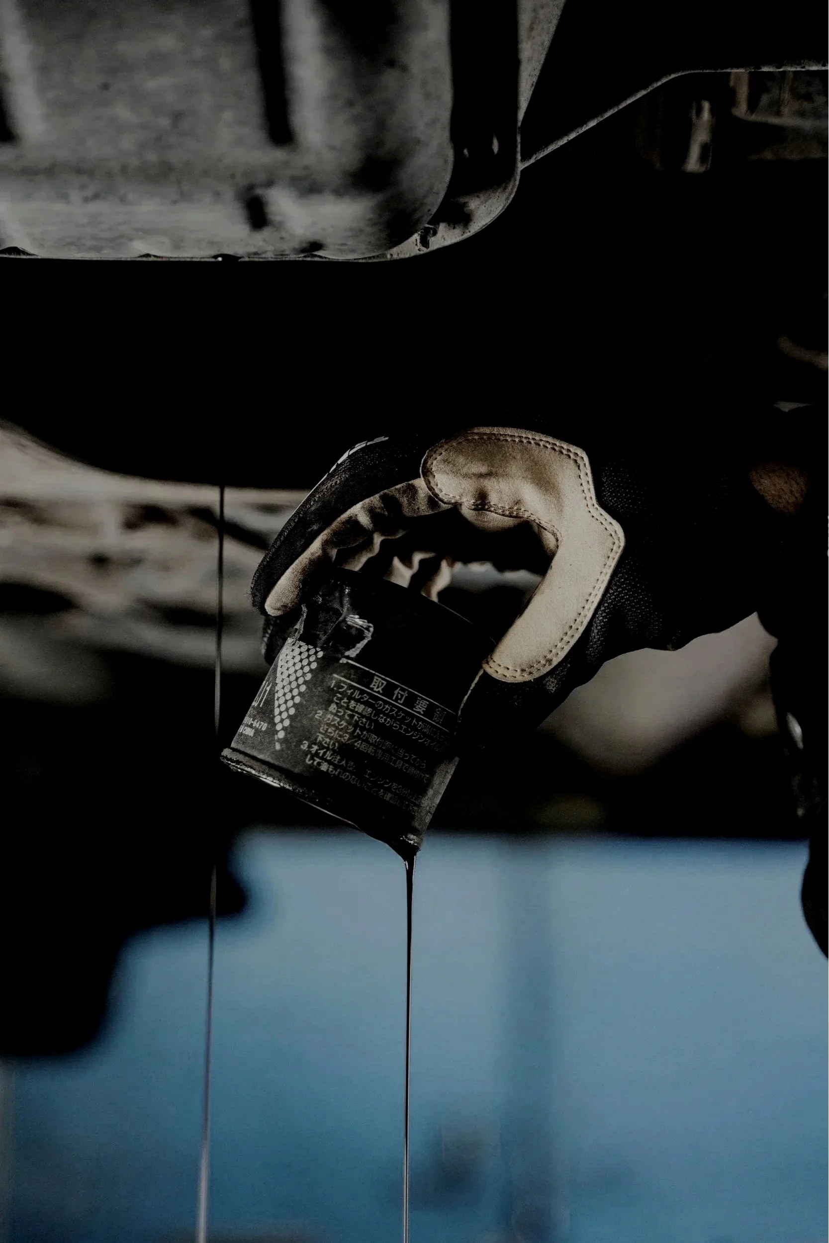 Close-up of a person wearing a glove holding a bottle of motor oil underneath a vehicle while oil drips from a drain plug.