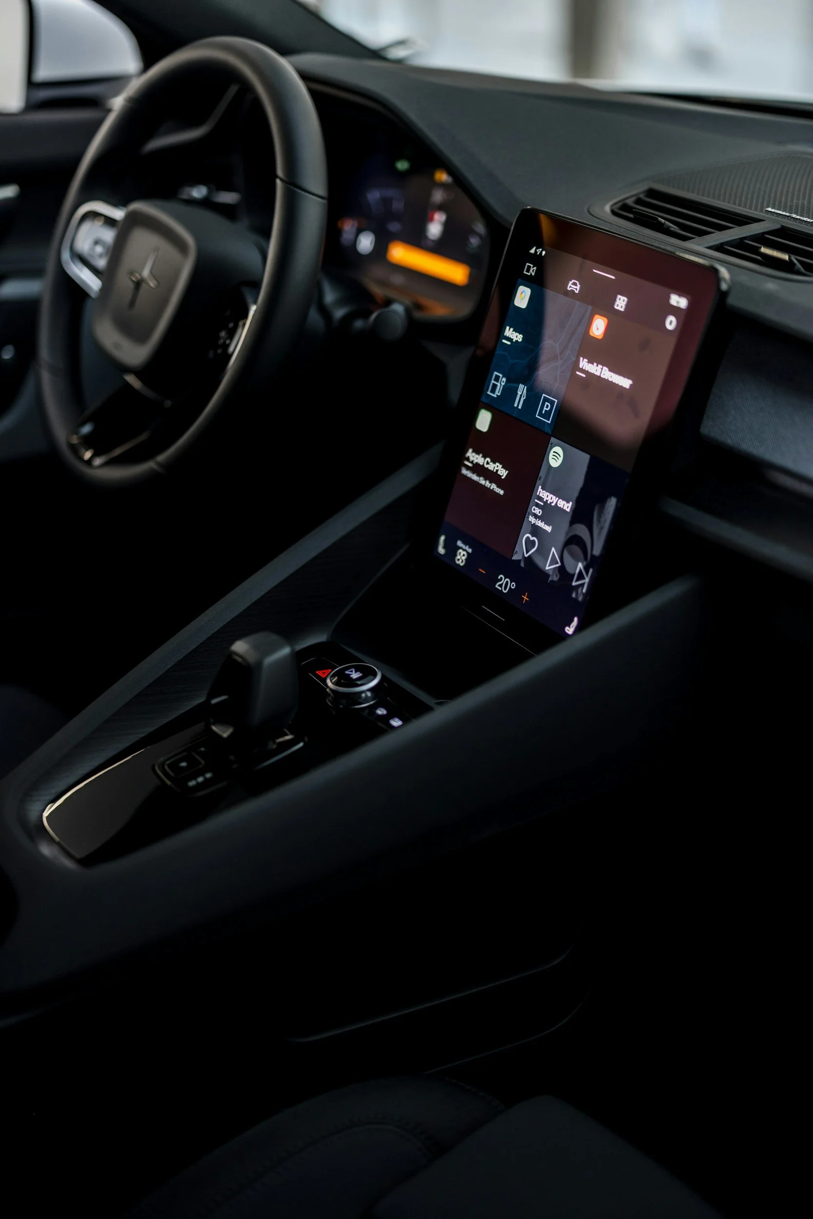 Inside view of a modern car dashboard showing the steering wheel with a logo, an infotainment screen displaying navigation and apps, and various control buttons. The interior is black with minimalistic design.