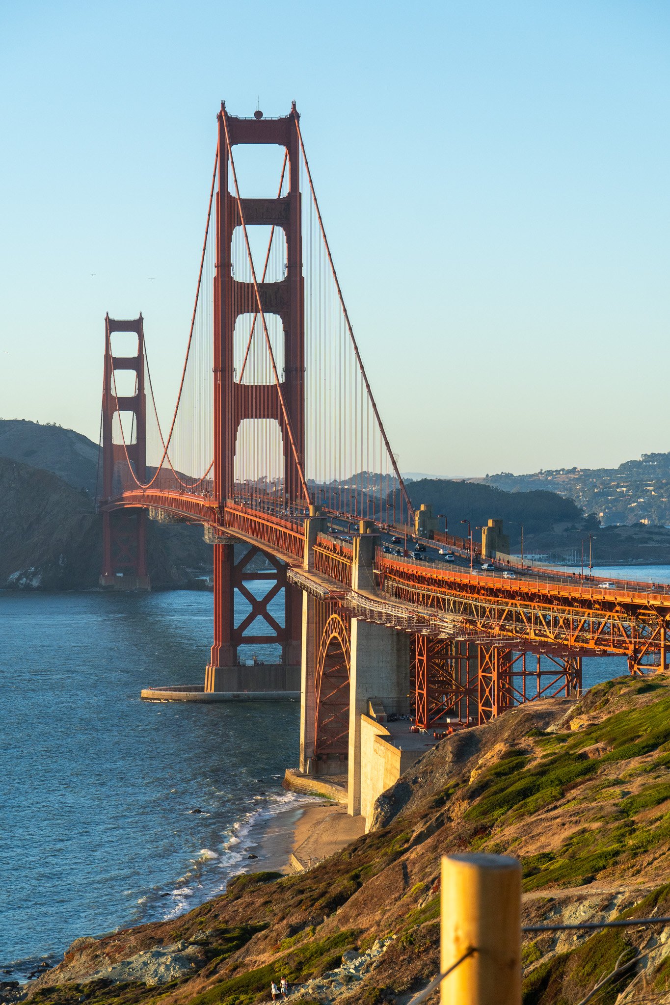 The Golden Gate Bridge in San Francisco, California, shown at sunset with pedestrians walking along a rocky, grassy shoreline in the foreground.