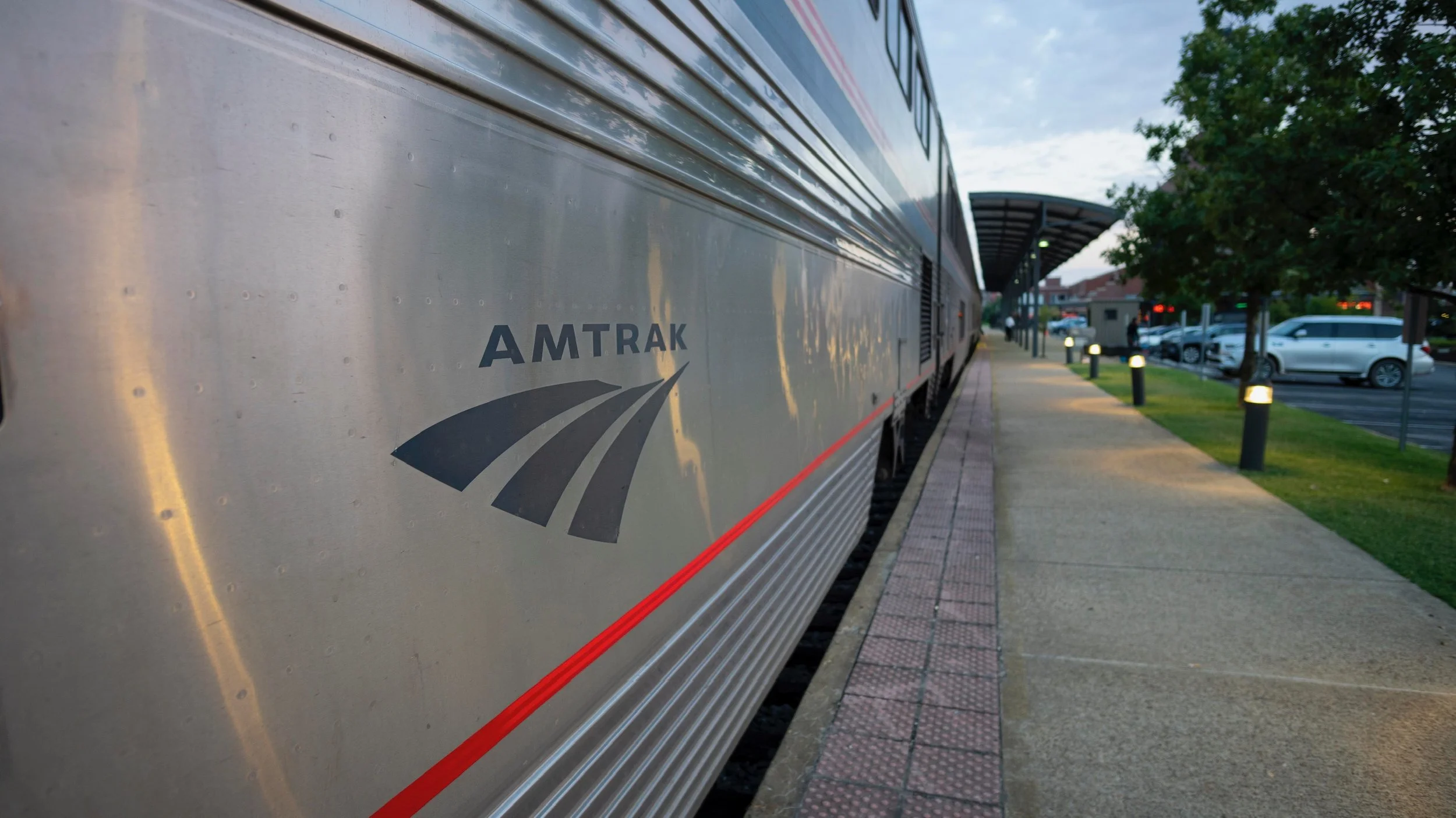 Amtrak train parked at a station platform during early evening with trees and parked cars in the background.