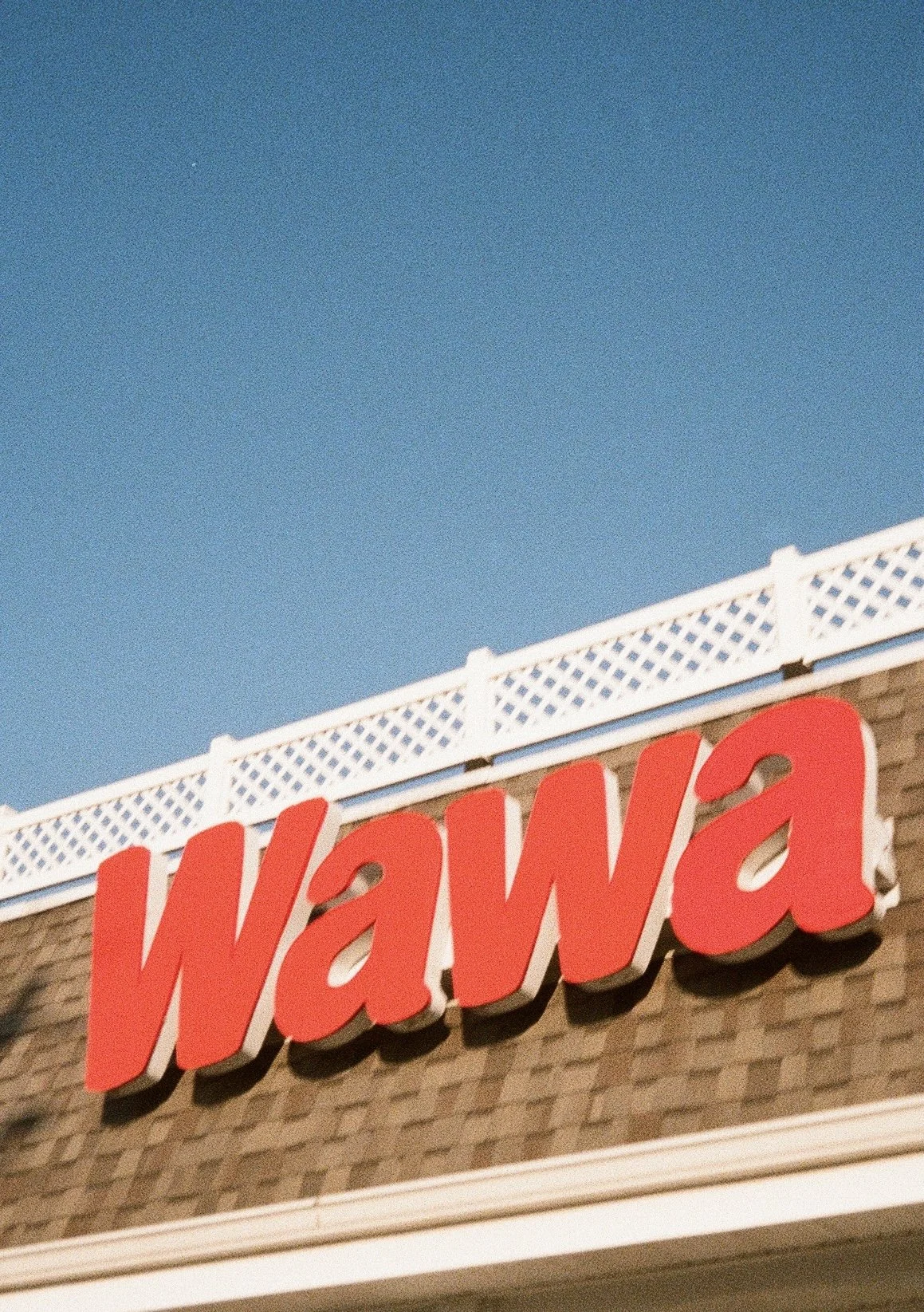 Close-up of a building sign with the word 'Wawa' in large red letters against a brick wall, with a white lattice fence and a clear blue sky in the background.