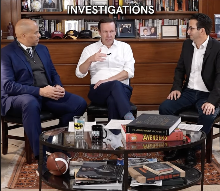 Three men sitting in a room decorated with books and memorabilia, engaged in a discussion, with a coffee table in front of them holding books, a glass with a Batman logo, a mug, and a football.