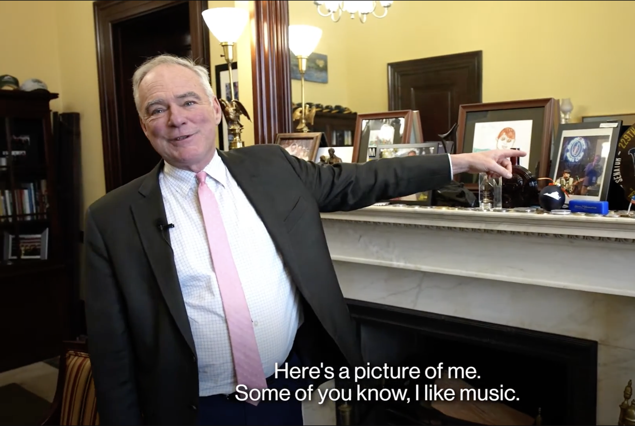 A man in a suit with a pink tie smiling and pointing to objects on a mantelpiece in a room with yellow walls and wooden furniture. The room has framed pictures and decorative lamps.