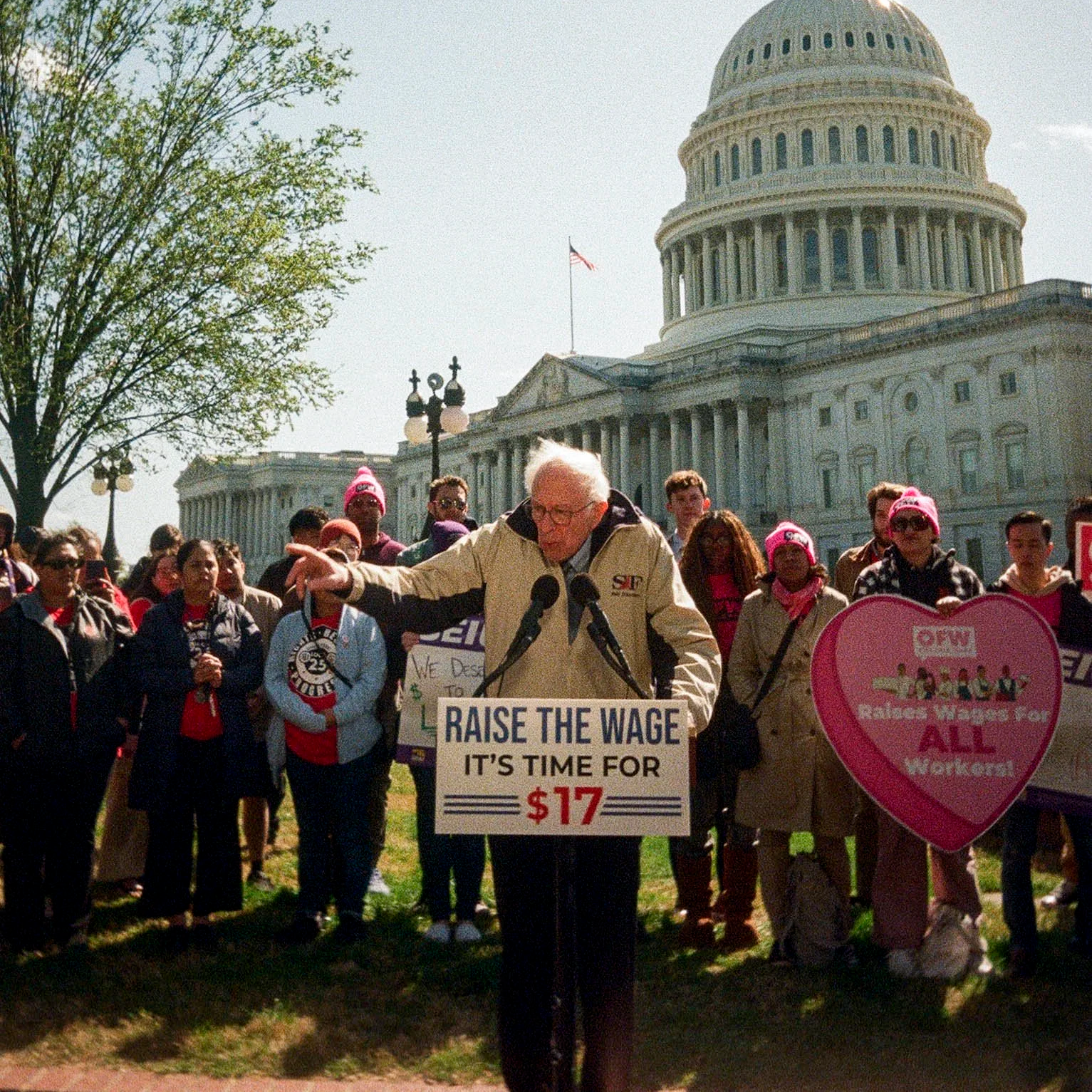  Senator Bernie Sanders speaking at a political rally outside the U.S. Capitol building, with a sign that says 'RAISE THE WAGE, IT'S TIME FOR $17'. 