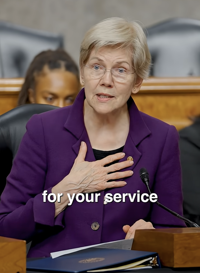An older woman with short blonde hair, wearing glasses and a purple blazer, speaks at a hearing with her right hand on her chest. A woman with braided hair and a dark outfit is visible in the background.