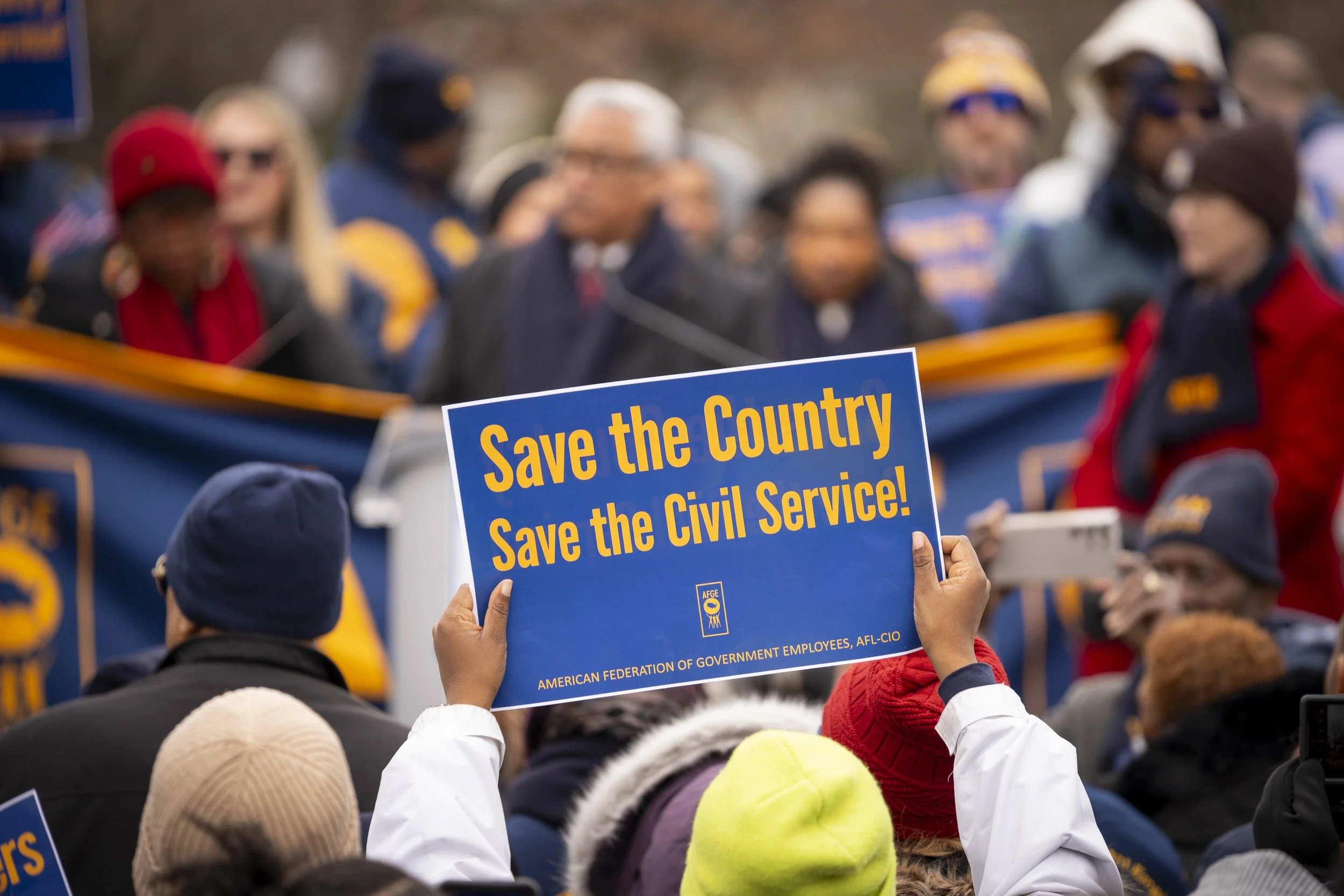 A crowd at a protest holding a blue sign that reads: 'Save the Country Save the Civil Service!' with a logo and additional text at the bottom.