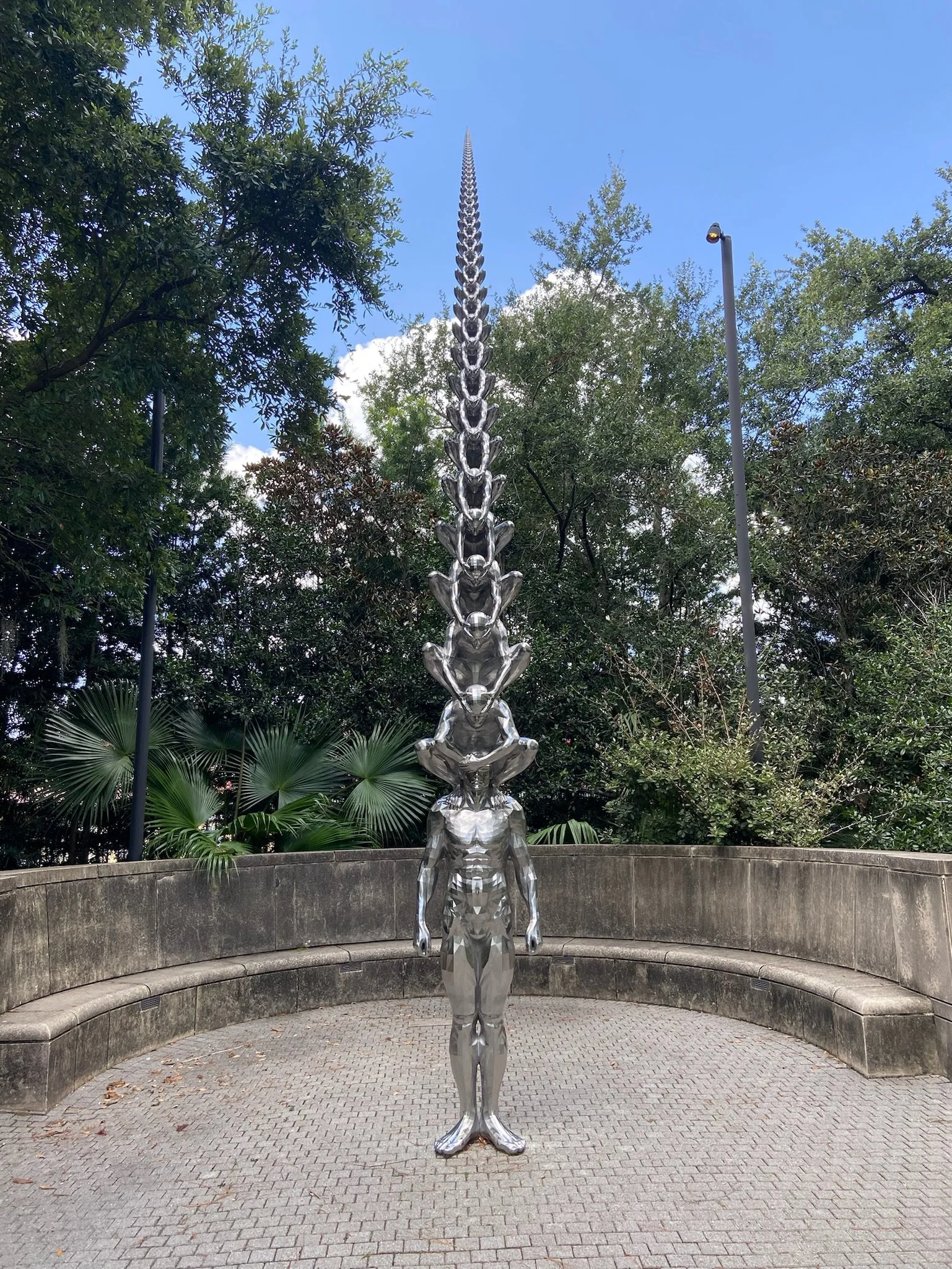 A silver metallic sculpture of a humanoid figure with ten frogs stacked on top of it, reaching a tall pointed tower, located in an outdoor park with trees and a stone bench surrounding it.