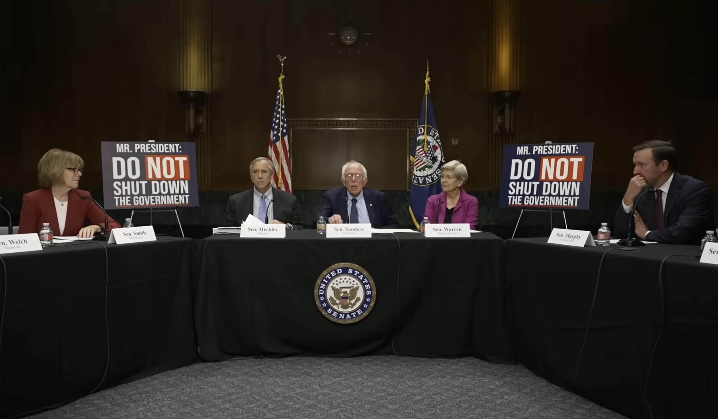 A group of five senators seated at a table during a Senate hearing or meeting, with signs that say 'Mr. President: Do Not Shut Down the Government.' The table has microphones, water bottles, and nameplates, and there are flags and a seal of the Unite
