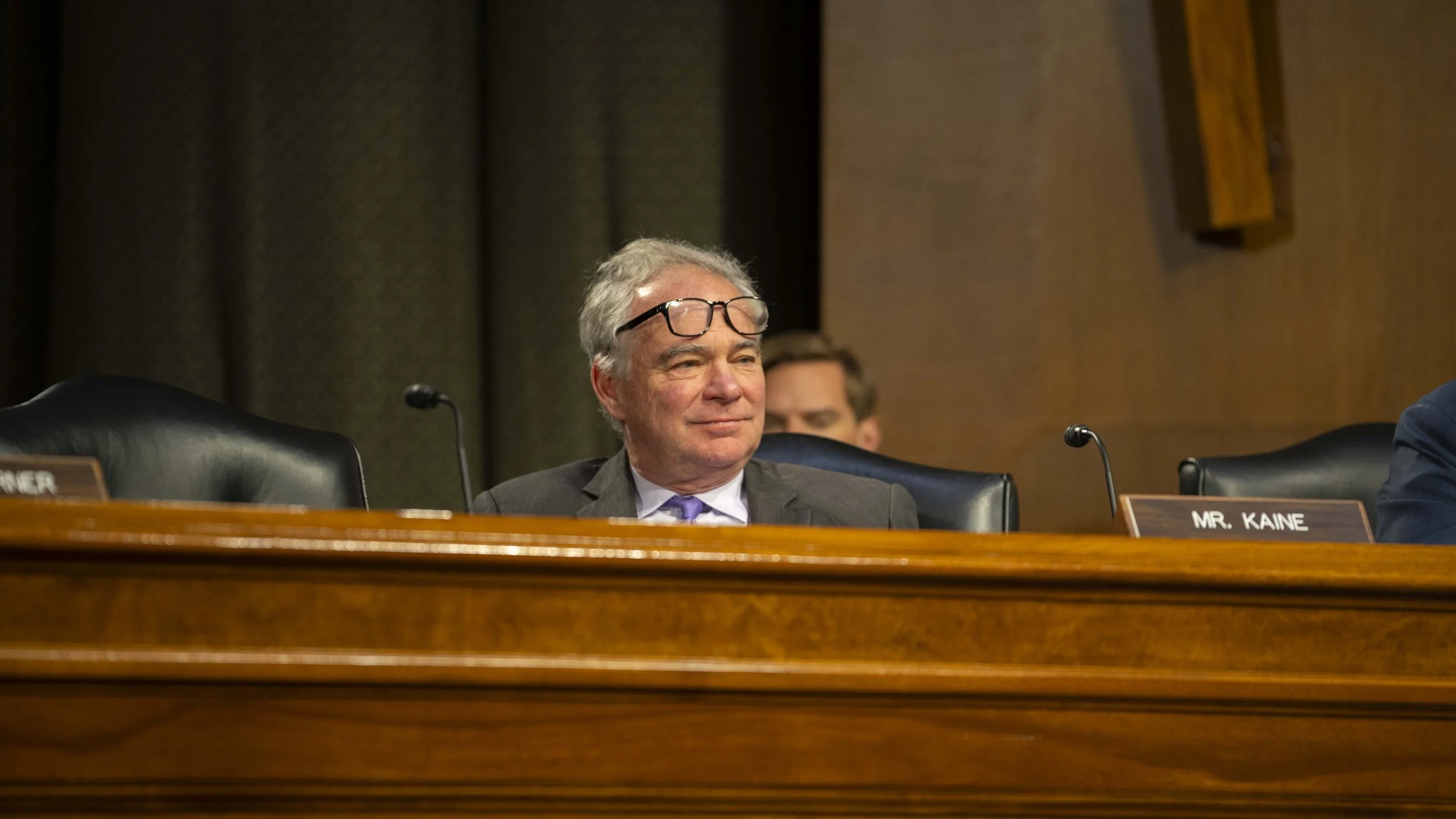 Senator Tim Kaine sits behind a committee room dais, his glasses on top of his head.