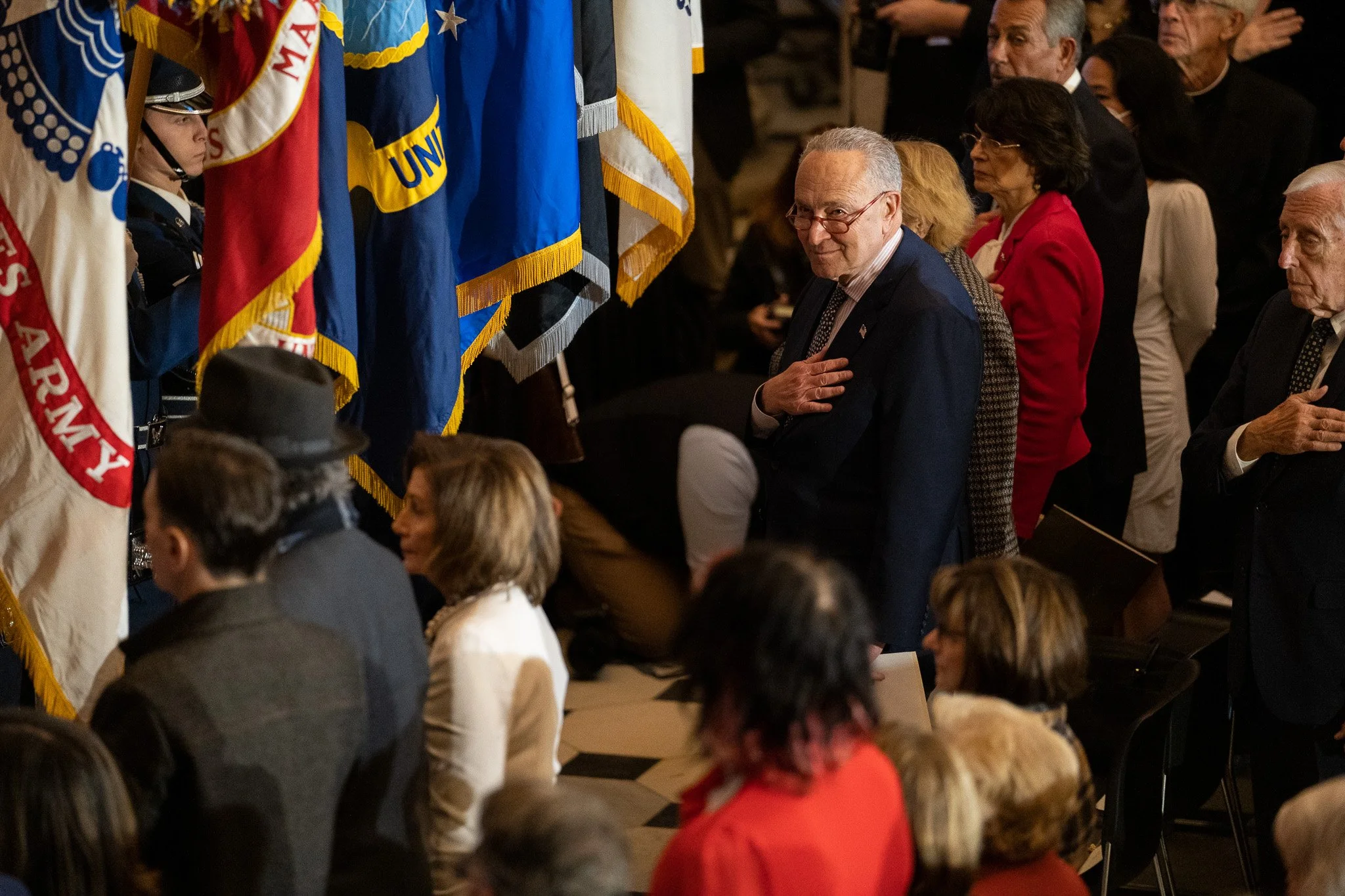 Sen. Chuck Schumer holds his hand over his heart as flags are carried past him in a crowd.