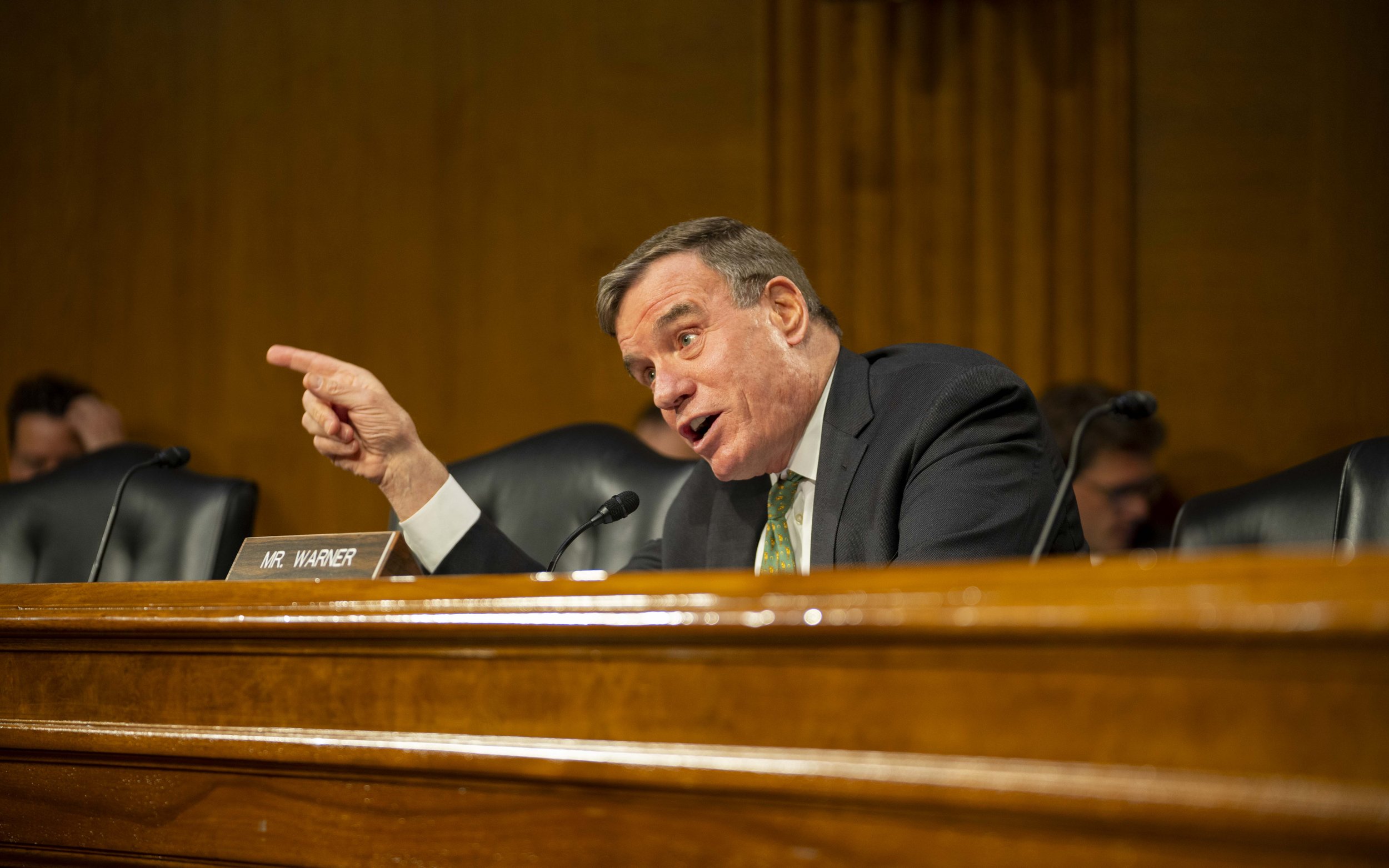 Senator Mark Warner speaking at a Senate hearing, gesturing with his hand and surrounded by microphones.