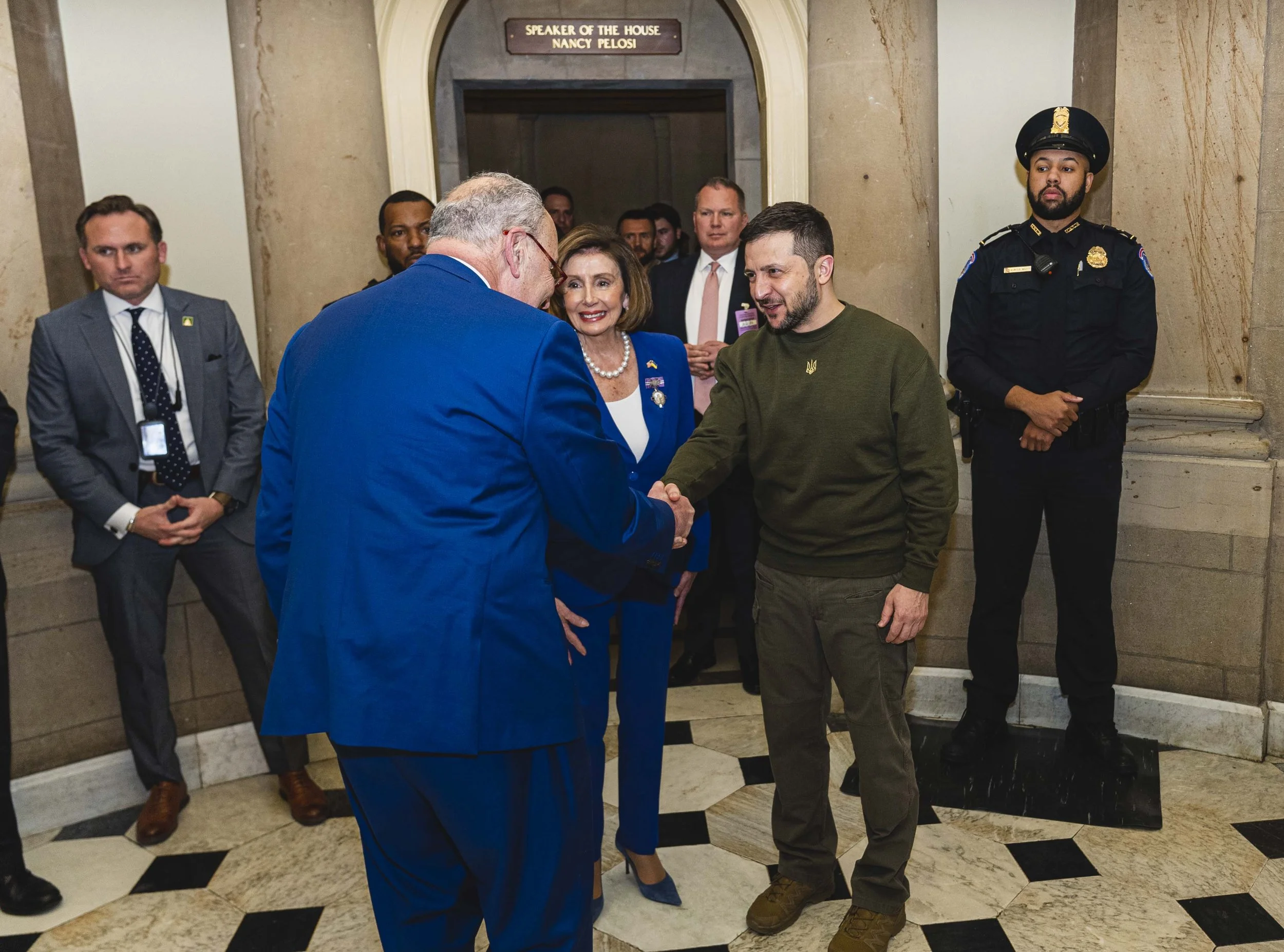 Chuck Schumer in a blue suit shaking hands with President Zelenskyy in a green shirt, with House Speaker Nancy Pelosi, surrounded by several security personnel and onlookers outside the Speaker's Lobby in the Capitol.