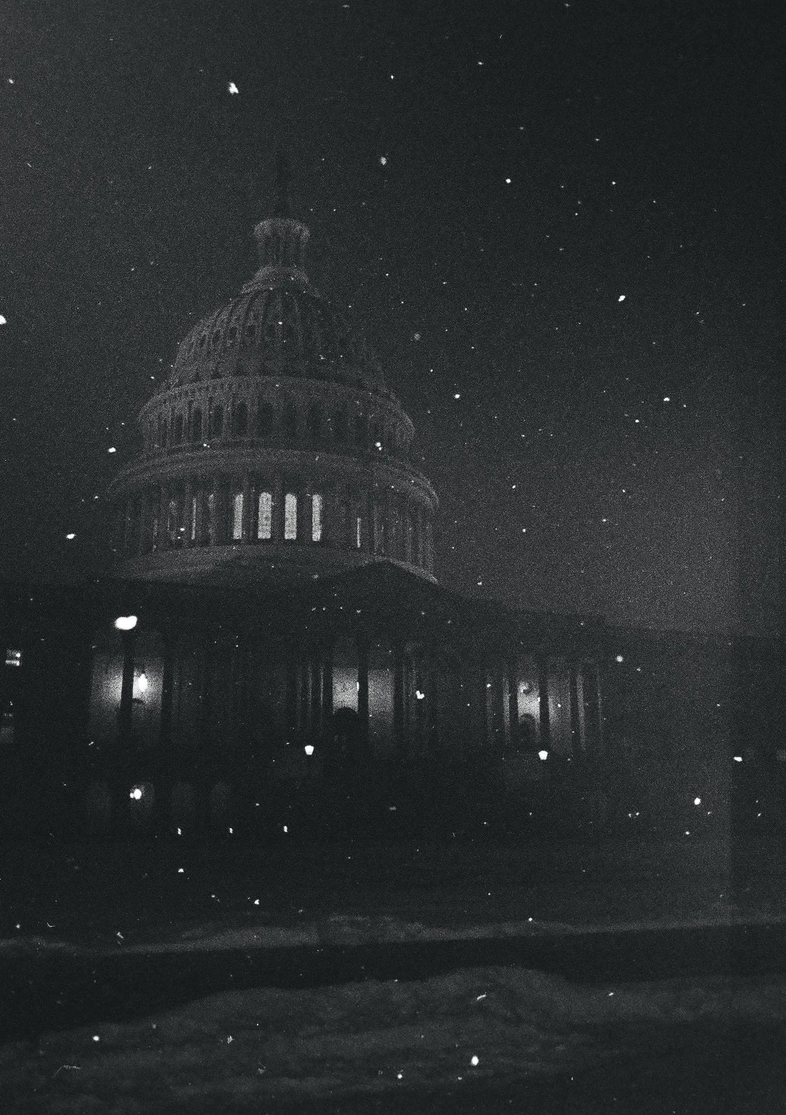 Nighttime scene of the U.S. Capitol building with snow falling and illuminated windows.